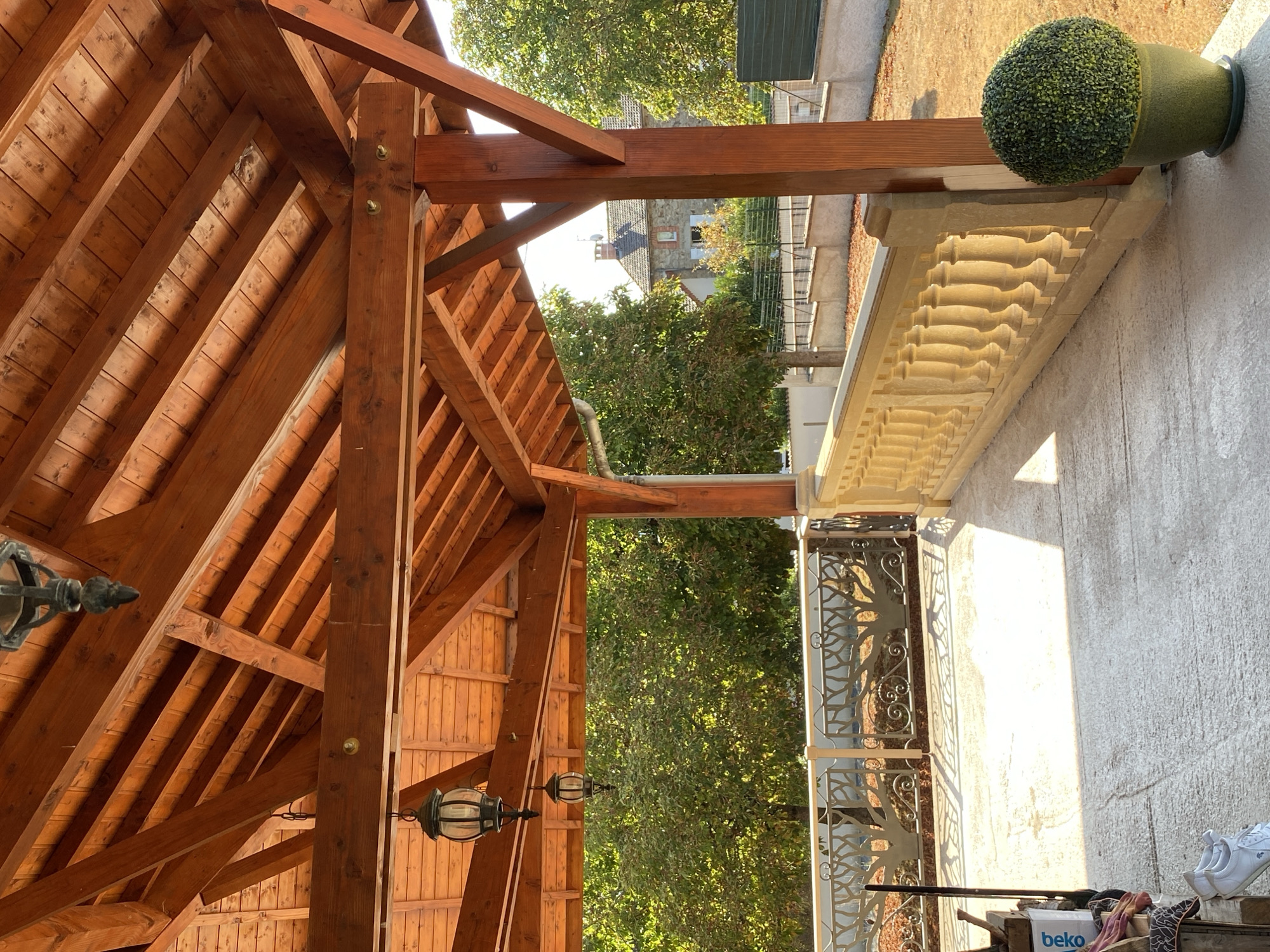 The image shows a covered outdoor patio area with a wooden roof structure. Two black lanterns hang from the wooden beams of the roof. A decorative metal railing with a tree motif runs along the edge of the patio, behind which is a stone balustrade. In the background, there are trees and a house with a tiled roof, suggesting a residential area in Courpière, France. Sunlight casts shadows across the concrete floor of the patio. A green potted plant is visible on the right side of the image. On the left side of the patio, a white pair of sneakers and some boxes are present.