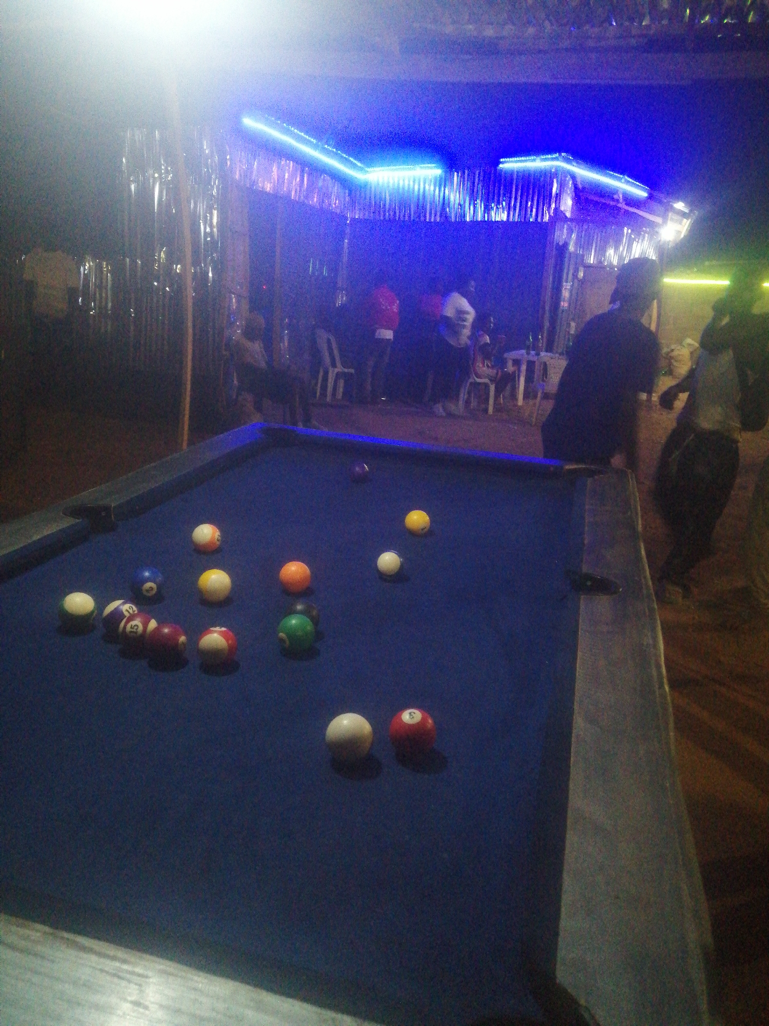 The image depicts a pool table with balls scattered on it, located in what appears to be an outdoor entertainment venue. The table is the central focus, with pool balls arranged haphazardly across its blue surface. Several individuals are visible in the background, some standing and others seated at tables. The venue is illuminated with blue and yellow lighting, suggesting a nighttime setting. The architecture appears to be of a casual or semi-permanent structure. Based on the prompt, this scene is located in Gura Top, Nigeria. The overall environment seems to be a social gathering spot.