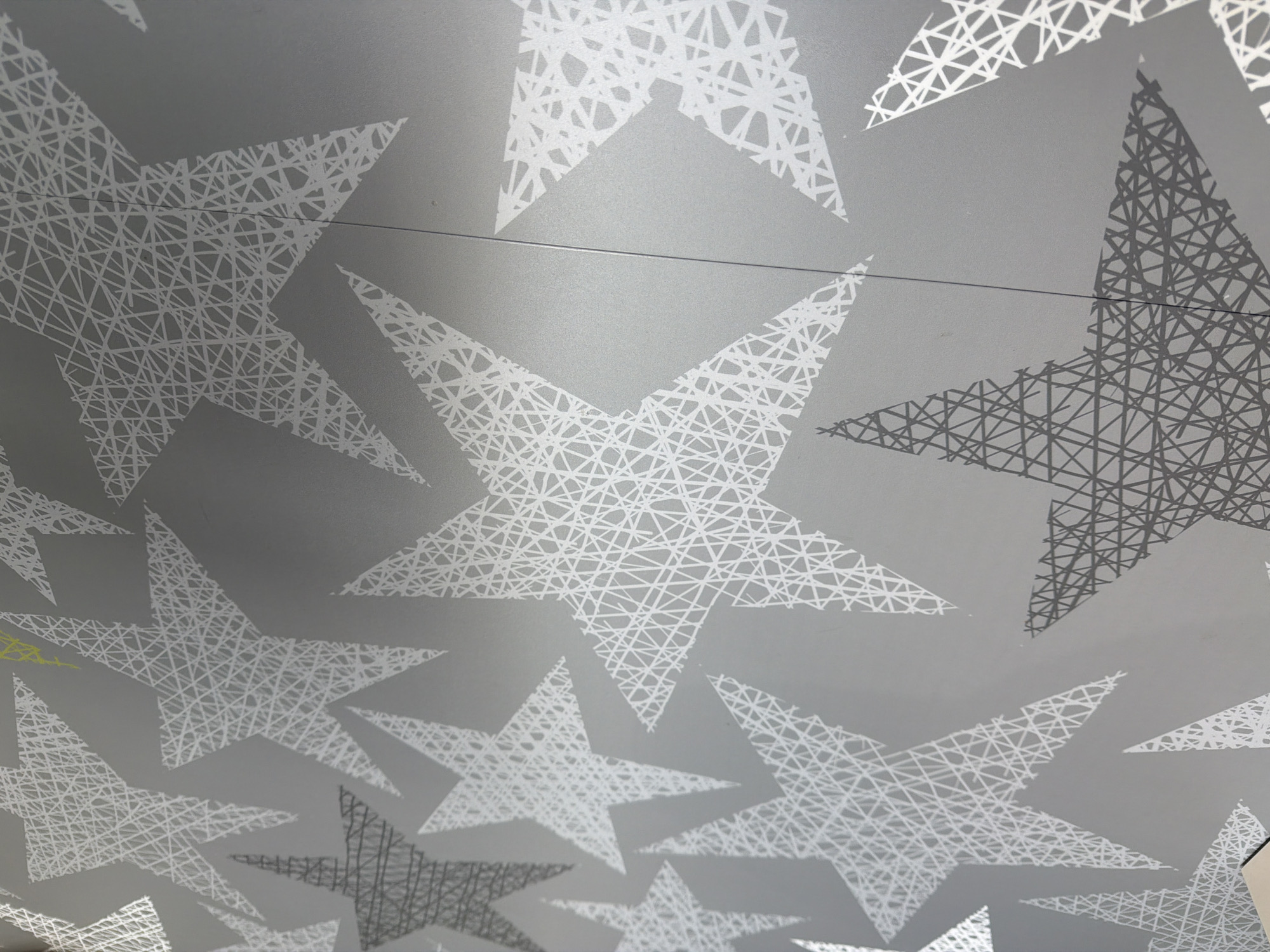 The image shows a close-up of a wall with a repeating pattern of abstract stars. The stars are outlined with a dense network of thin, white lines on a grey background. Some stars appear as solid grey shapes, suggesting a shadow or a different print. The wall has a subtle texture. There is a faint vertical line running down the center-right of the image, indicating a seam between two panels or sections of the wall. There are no people or objects visible in the image, nor any clear indication of the specific location within Kyiv, Ukraine.