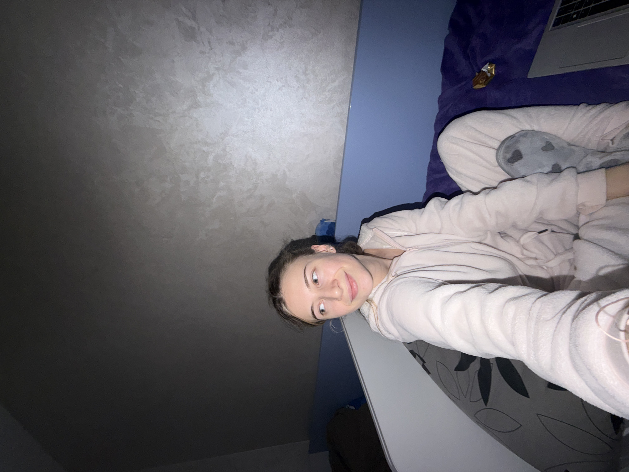 A woman with brown hair is lying in bed, looking at the camera. She is wearing a light pink hooded jacket and patterned grey socks. A laptop rests on a dark blue blanket next to her. There is a piece of chocolate on the blanket next to the laptop. A wall is visible in the background. The image was not taken in Plovdiv, Bulgaria, as I lack the information to confirm the location.