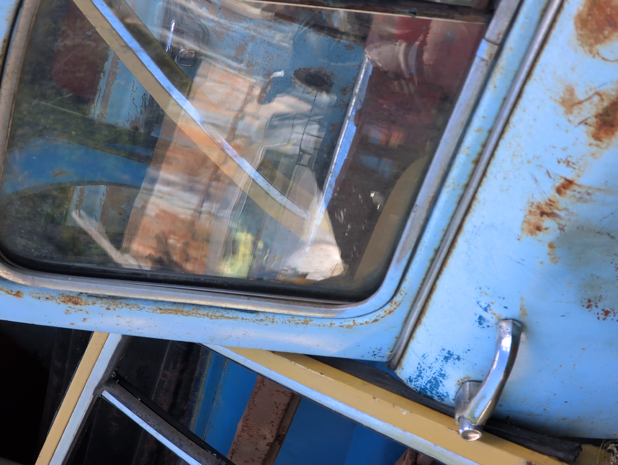 The image is a close-up of a light blue, heavily rusted vintage vehicle, likely a car or van. The focus is on the side door and window, showing the weathered paint, visible rust patches, and a chrome door handle. The glass window is dirty and reflects the surroundings, which appear to be an outdoor setting with other vehicles or structures. There are no people or animals visible in the frame. The scene is suggestive of a junkyard or an abandoned vehicle lot. The location is identified as Novoe Devyatkino, Russia.