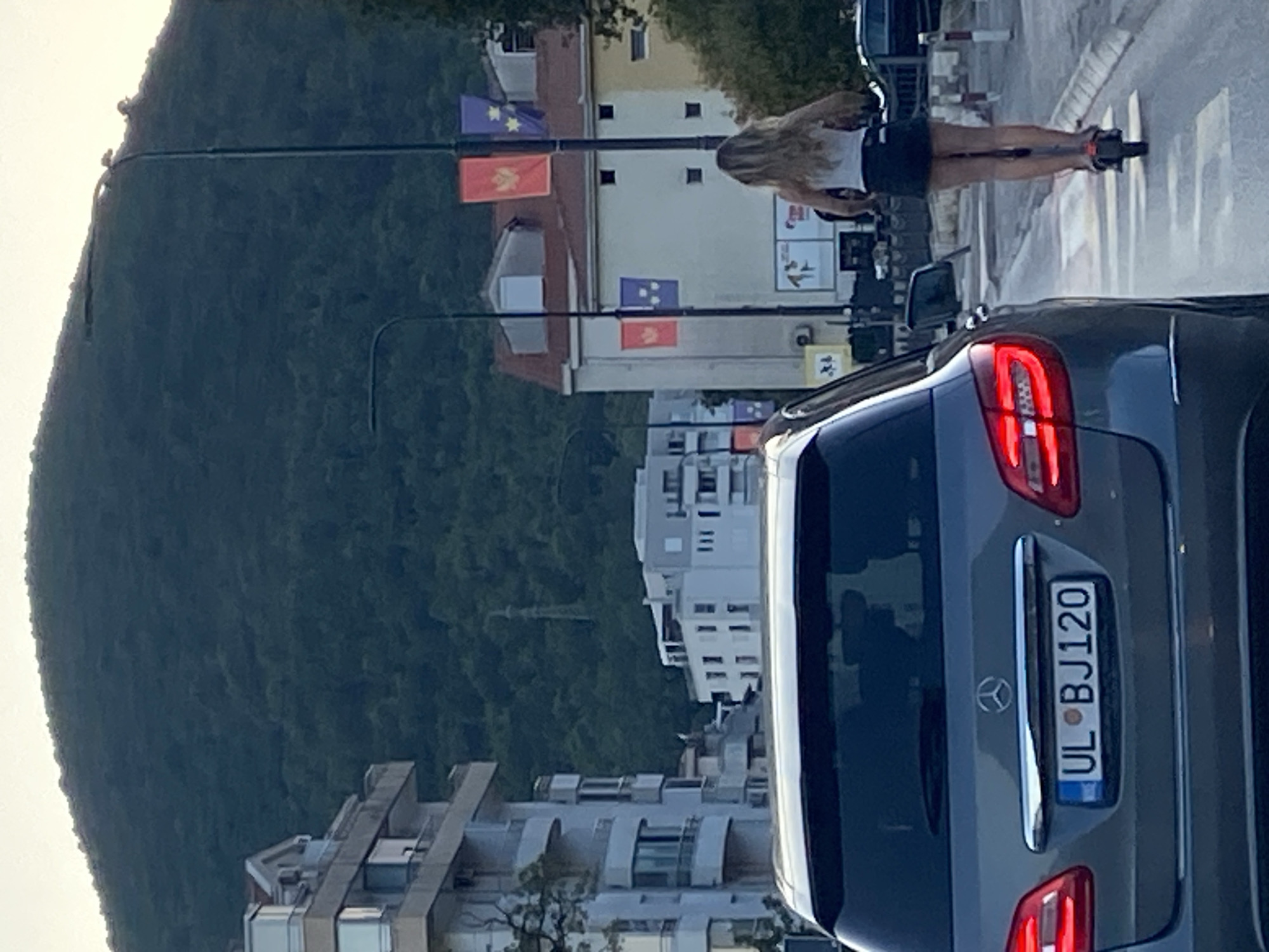 In Budva, Montenegro, a woman with blonde hair rides an electric scooter down a street. She is wearing a white tank top and a dark skirt. In the foreground, the rear of a grey Mercedes-Benz sedan is visible, displaying a license plate that reads "UL BJ120". Behind the woman, a building with red and purple flags hanging from lampposts is seen. In the background, a lush green mountain overlooks the city, with a distant power line tower visible on its slope. The scene appears to be set during daylight hours, with the sky a pale color.