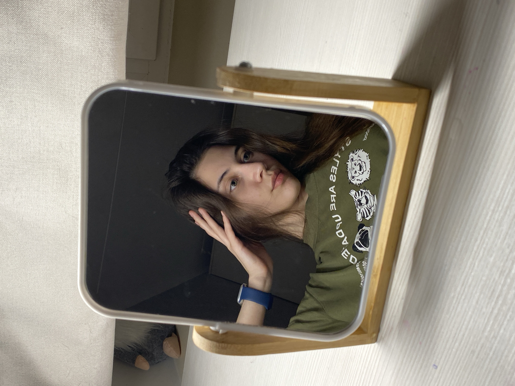 A young woman is seen in a mirror, reflected as if she is in a room or space beyond the mirror’s surface. She has long brown hair and wears a green t-shirt with white print and cartoon-like designs. On her wrist is a blue smartwatch. She is touching her hair with her left hand. The mirror has a light wood stand. The background of the surrounding room or space is ambiguous, with white walls and furniture partially visible, including a stuffed animal. There is no information given to identify a location in Votkinsk, Russia.