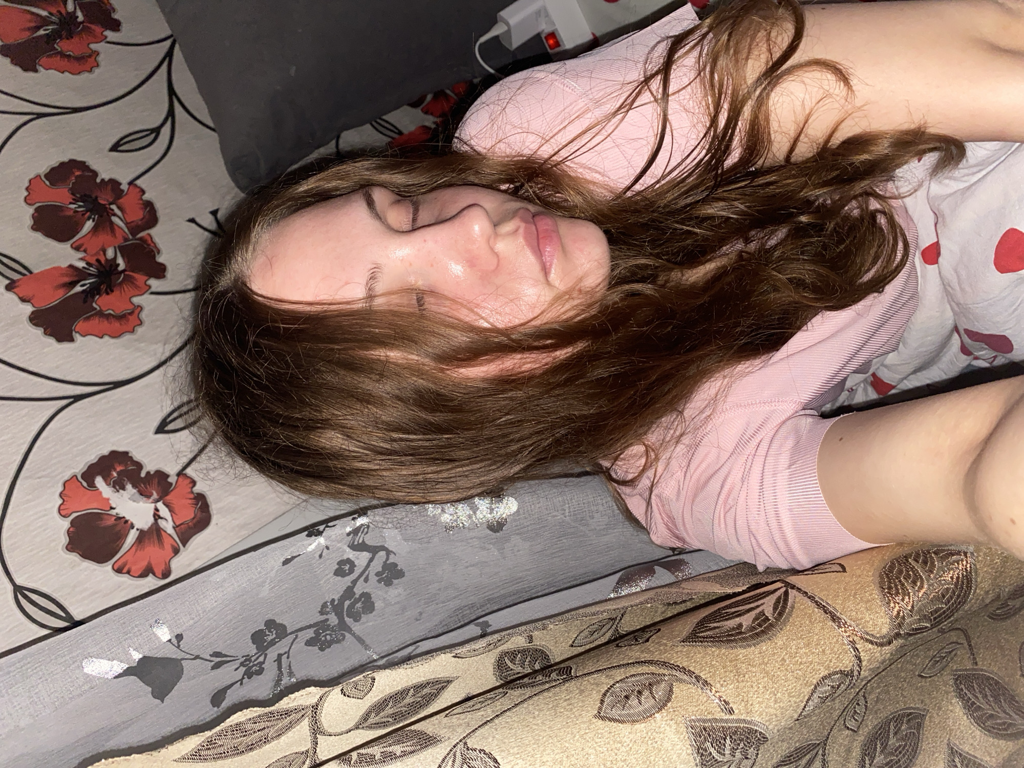 The image shows a close-up, overhead view of a young woman lying on her side with her eyes closed, seemingly asleep or resting. She has long, wavy brown hair that falls around her face and shoulders. She is wearing a pink ribbed t-shirt. Her face is slightly flushed, and her skin appears dewy. The background consists of patterned wallpaper with floral motifs and draped curtains with leaf patterns. A pillow is visible behind her head, and a white power strip with red indicator lights is partially visible to her right. The scene appears to be indoors, possibly in a bedroom. There is no information available about the location in the city of Вассята, Russia, as the image itself does not contain any identifying geographical markers.