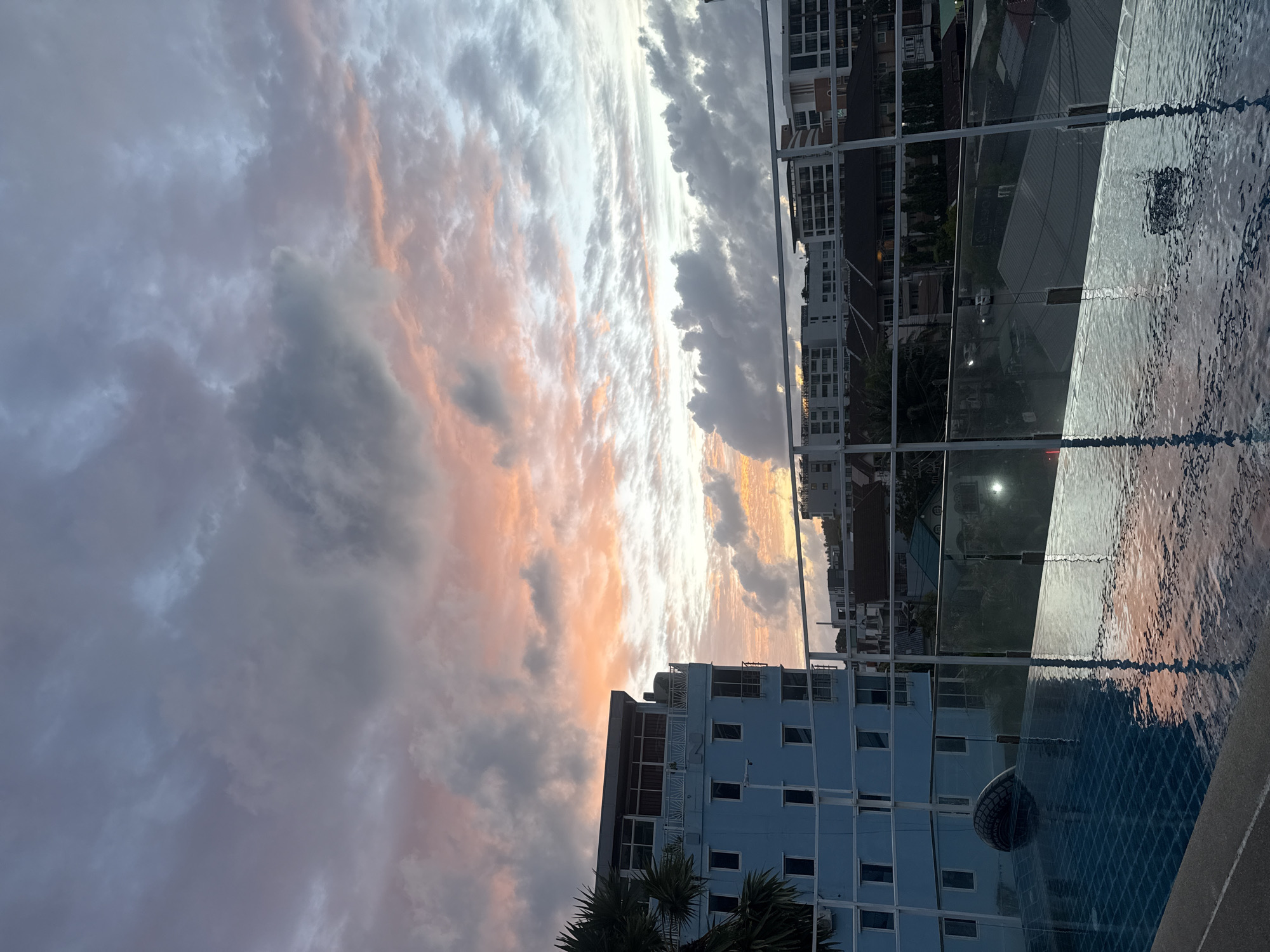 The image captures a rooftop swimming pool overlooking the city of Karon, Thailand, at dusk. The sky is a canvas of dramatic clouds, with hues of pink, orange, and grey suggesting the last light of the setting sun. A light blue building, possibly a hotel, stands prominently on the left, its windows reflecting the sky. In the foreground, the rippling surface of the swimming pool mirrors the colorful sky and the surrounding buildings. The reflection creates a sense of depth and tranquility. Beyond the pool, a low-rise cityscape is visible, characterized by residential buildings and lush tropical vegetation. The overall scene evokes a sense of peacefulness and the beauty of a tropical sunset.