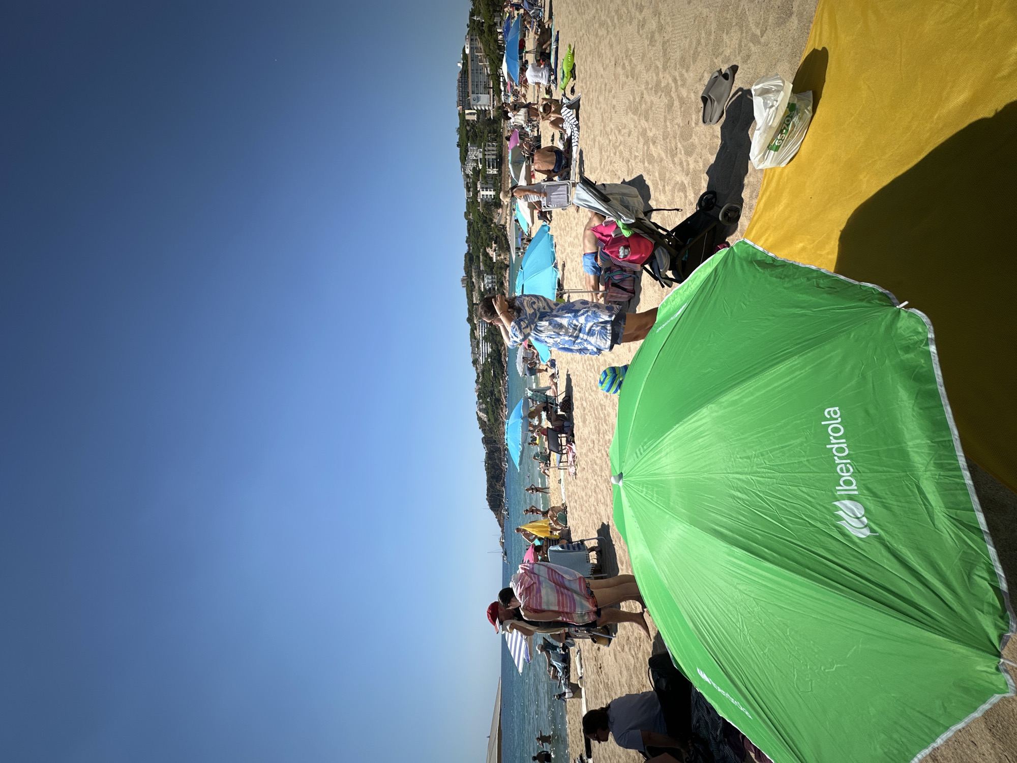 The image depicts a crowded beach scene in Sant Feliu de Guíxols, Spain, on a sunny day. The foreground is dominated by a large, bright green umbrella with the "Iberdrola" logo visible, casting a shadow over a yellow beach towel. Beyond this, the sandy beach is populated with numerous people lounging under umbrellas, sitting on chairs, and walking. In the mid-ground, several brightly colored umbrellas, including blue ones, provide shade for beachgoers. Some individuals are partially submerged in the calm, blue sea in the background, with a few sailboats anchored offshore. A line of buildings and lush green vegetation covers the hillside overlooking the beach. The sky is a clear, cloudless blue.