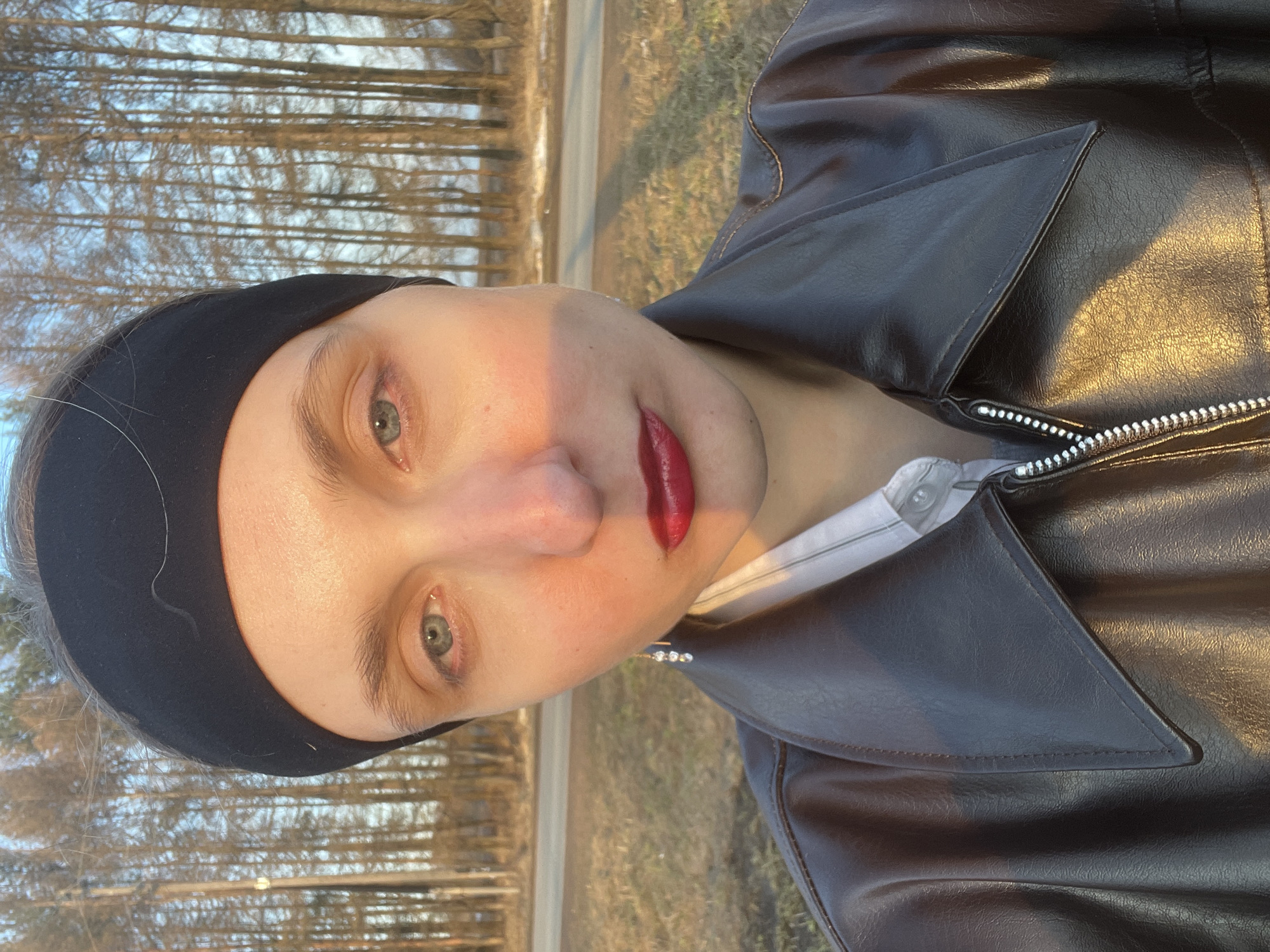 A close-up selfie shows a person wearing a black headband and a dark leather jacket. A light-colored button-down shirt is visible beneath the jacket. The person has light skin, green eyes, and dark red lipstick. The background includes trees and what appears to be a road or path. It is daytime, with sunlight hitting the subject's face. Without additional information or landmarks, precise location in Подшивалово, Russia cannot be determined.