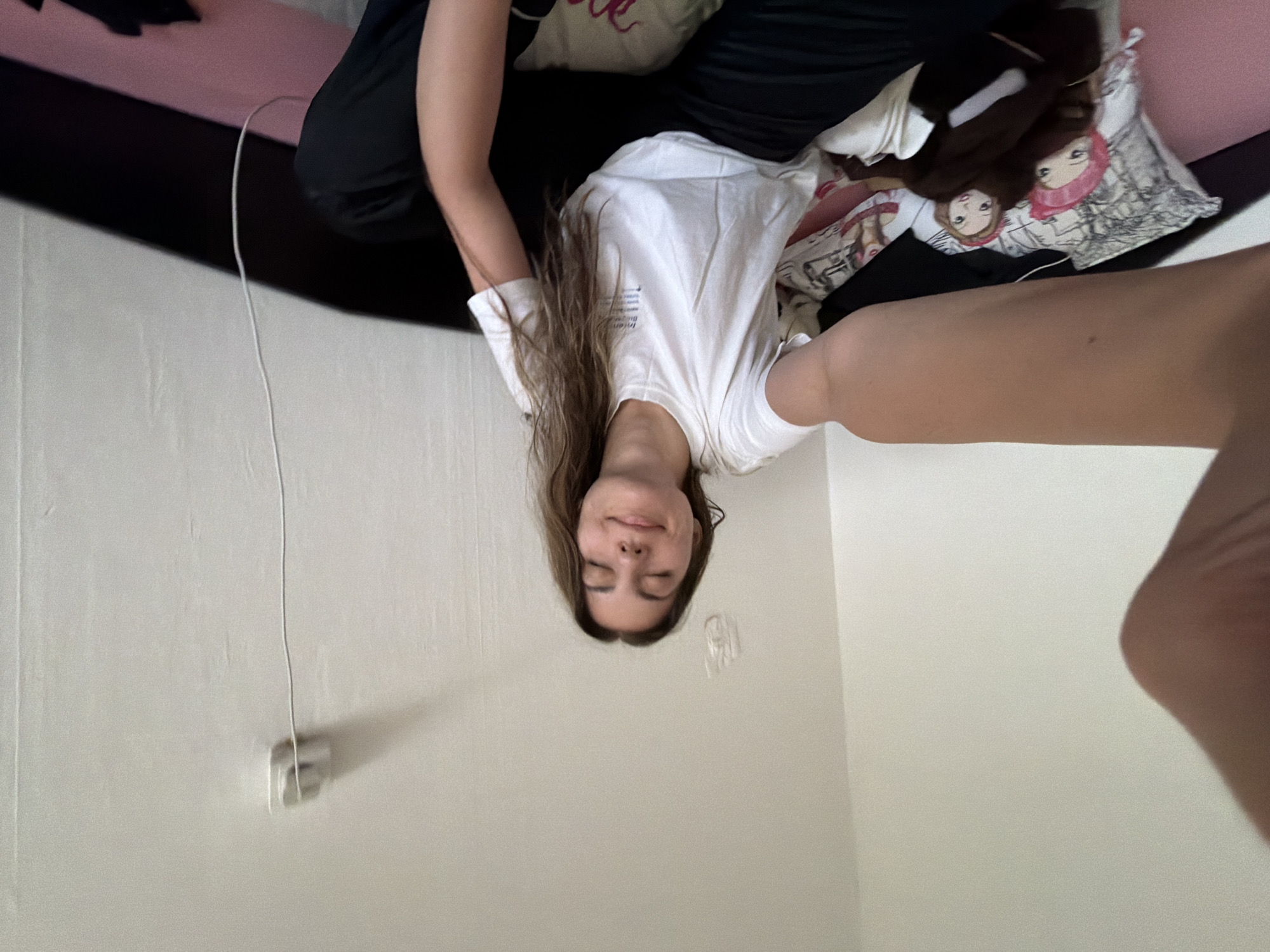 A person is lying on a bed with a pink sheet, pillows with cartoon characters and text, and black clothing. They are wearing a white t-shirt and their long brown hair is hanging down. The person's face is visible. A white wall with an electrical outlet is in the background. The location cannot be determined from the image.