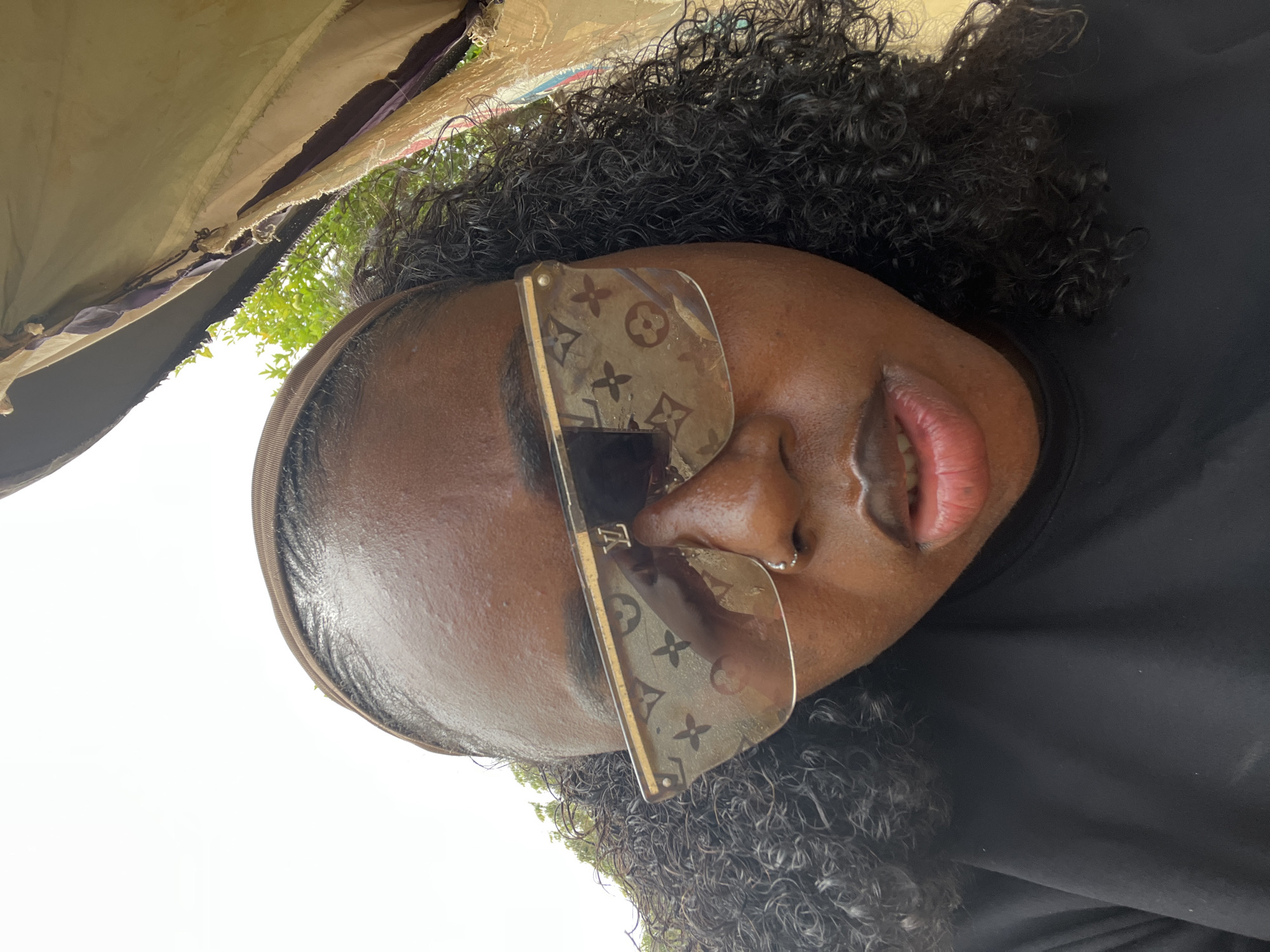 A close-up, low-angle, outdoor selfie shows a Black woman with curly dark hair, wearing a brown headband and large, clear sunglasses with a Louis Vuitton monogram print. She is also wearing a black top and a silver nose ring. Her lips are slightly parted. The background is slightly blurred, showing some greenery and what appears to be a makeshift tent or shelter with torn fabric. The scene appears to be in an outdoor setting, possibly a market or a casual outdoor gathering space in Abuja, Nigeria. The lighting suggests it is daytime.