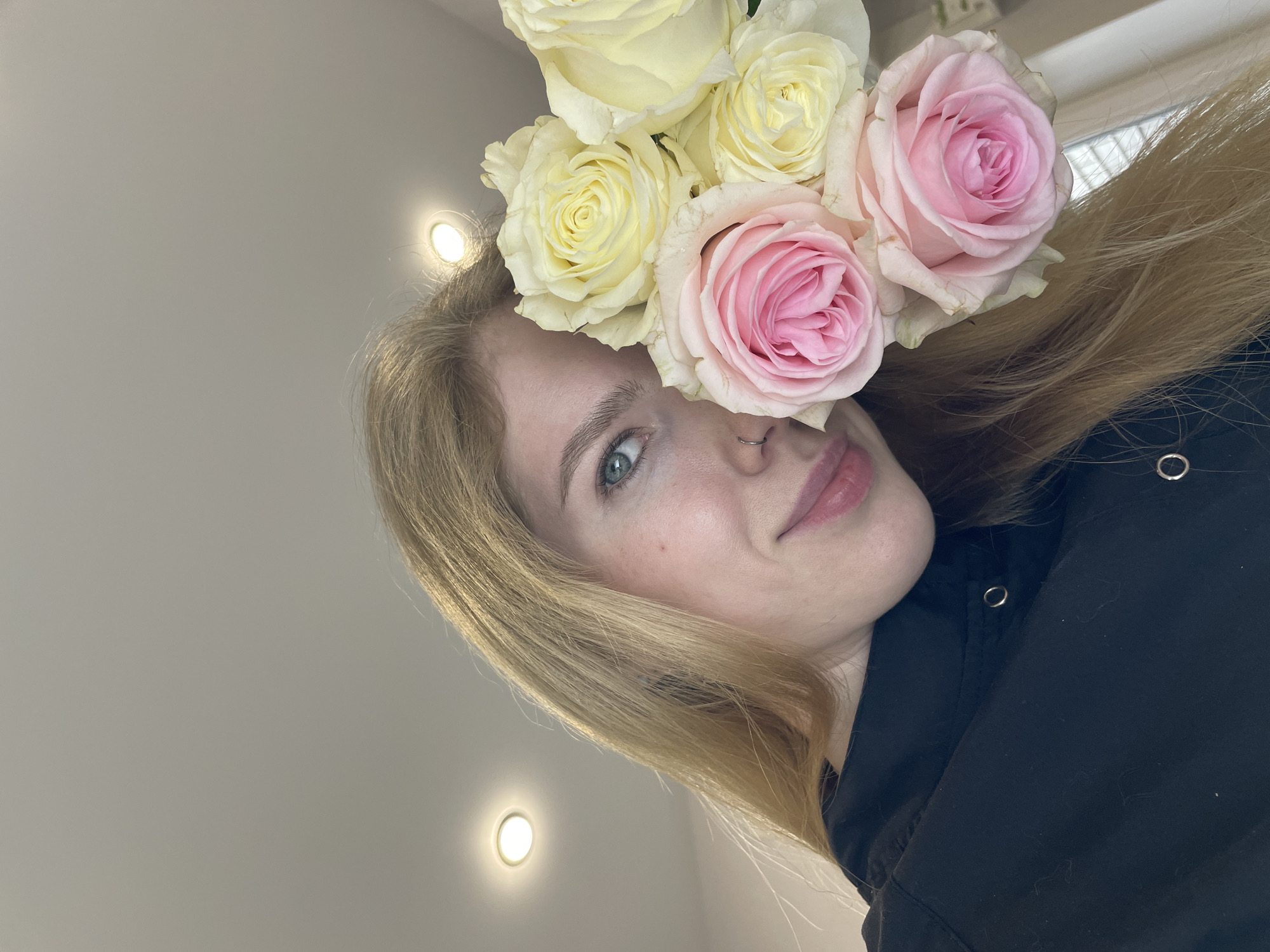 A low-angle, close-up portrait captures a young woman indoors, likely in Izhevsk, Russia, looking upwards and to the right with a soft, gentle expression. She has light blonde hair that cascades around her face and over her shoulders, and clear, light-colored eyes. Several roses, including pale yellow and soft pink varieties, are artfully arranged on top of her head, nestled in her hair, almost like a floral crown. A small silver nose piercing is visible on her left nostril. She is wearing a black garment, possibly a hoodie or jacket, with metallic eyelet details visible on the fabric near her shoulder. The background is a plain, light-colored (likely white or light grey) ceiling with two recessed spotlights emitting a warm, circular glow. No text is visible in the image.