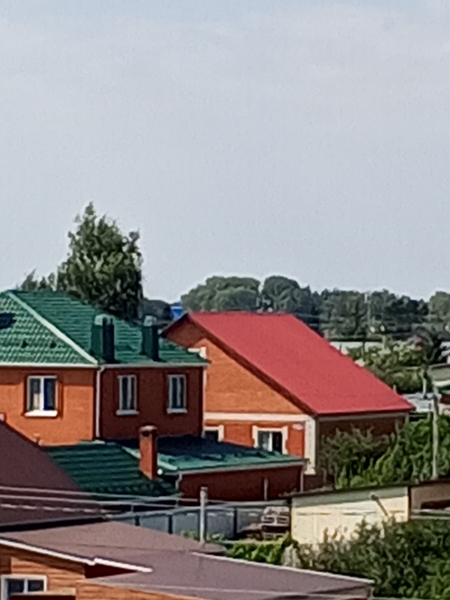 The image depicts a suburban residential area with several houses under a bright, overcast sky. The foreground shows the angled roofs of several houses, one with a dark brown, shingled roof and another with a lighter wooden siding and a dark brown roof. In the mid-ground, there are two brick houses. The house on the left has a green, corrugated metal roof and orange brick walls with white-trimmed windows. The house on the right has a bright red, corrugated metal roof and orange brick walls with white-trimmed windows. Behind these houses, more residential buildings and trees are visible, suggesting a densely populated area. The overall impression is one of a typical Russian dacha settlement, likely located on the outskirts of a city like Chistopol, Tatarstan, Russia, given the architectural style and prevalence of detached homes with individual gardens or plots. The scene is static, with no people or animals visible, and no immediate action occurring. The environment appears to be a sunny day, despite the overcast sky, indicated by the shadows cast on the buildings.