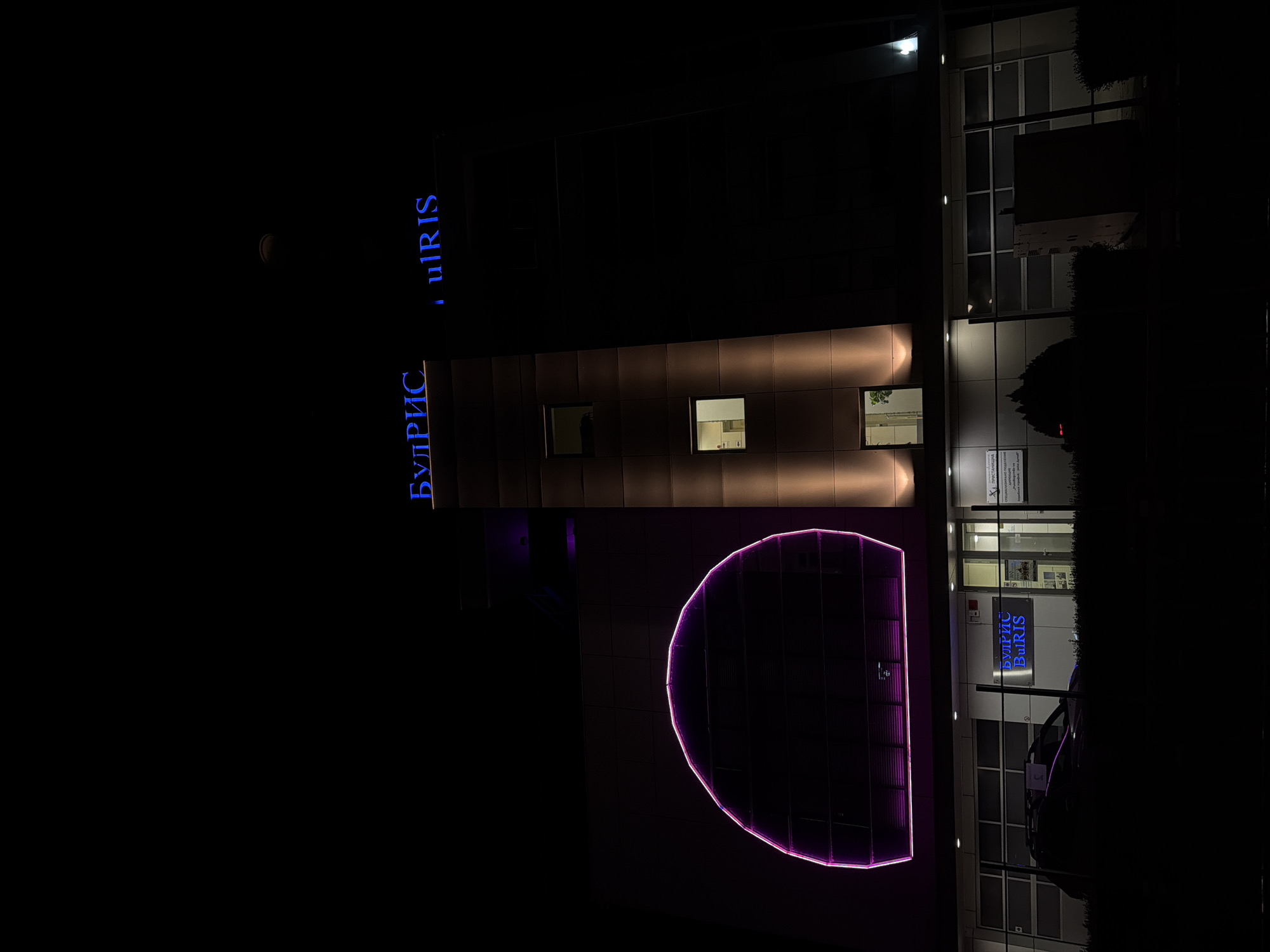 The image shows a modern building at night in Ruse, Bulgaria, with illuminated signage and architectural features. The building has a sleek, dark facade with segmented panels. Prominently displayed on the upper part of the building is a neon blue sign that reads "БУЛРИС" and "ulRIS". To the left, a large, semi-circular neon pink light installation is affixed to the building's exterior.
On the lower level of the building, there is another illuminated sign that reads "БУЛРИС" in blue neon. A car is parked in front of the entrance, partially obscured by shadow. Various windows are visible, some illuminated from within, suggesting occupancy. The ground in front of the building is covered with dark vegetation. The overall impression is one of a contemporary business or office building, likely illuminated for nighttime visibility and aesthetic appeal. The presence of Cyrillic script on the signage indicates the location is in Bulgaria.