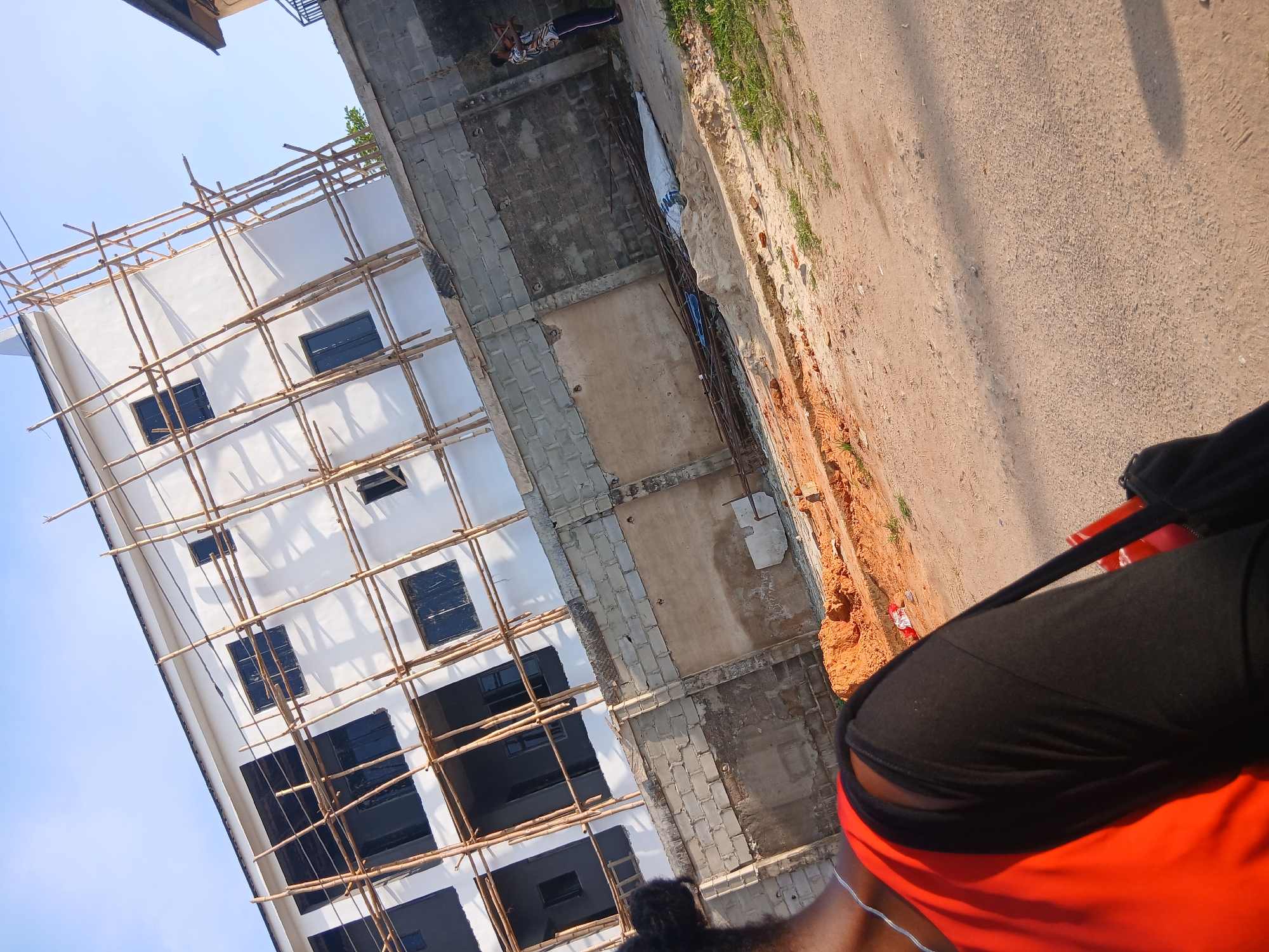 A low-angle shot displays a building under construction in Lagos, Nigeria. The building, partially constructed, has a white facade with black window openings. A wooden scaffolding surrounds the building. Adjacent to the building is an unfinished section with concrete blocks and exposed walls. A dirt-covered patch separates the building from a paved road. The lower right corner of the frame features a person with an orange shirt and black jacket.