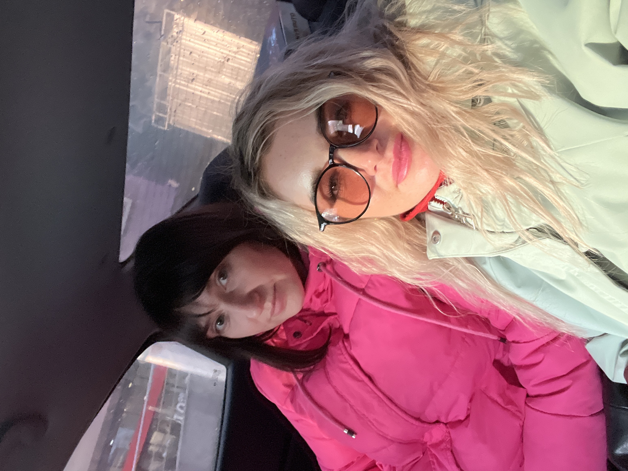 The media file displays two women inside a vehicle, likely a car or bus, in Izhevsk, Russia. The image appears to be a selfie, with the subjects positioned closely.
On the right, a woman with long, wavy blonde hair is wearing prominent round, amber-tinted sunglasses with dark frames. She has pink lipstick and is dressed in a light-colored, possibly mint-green or pale grey, jacket. A red strap or collar detail is visible near her neck. Her expression is neutral.
Leaning in from the left, a woman with dark hair and bangs wears a bright pink puffer jacket with visible drawstrings. She has a subtle, gentle smile and is looking slightly upwards.
The background seen through the vehicle's window is blurred and shows reflections, along with what appear to be raindrops on the glass, suggesting rainy or wet weather outdoors. Faint outlines of buildings or structures are visible through the window. The interior of the vehicle, particularly on the left side of the frame, is dark. The lighting is soft, possibly indicating an overcast day or interior light. No discernible text is visible in the image.