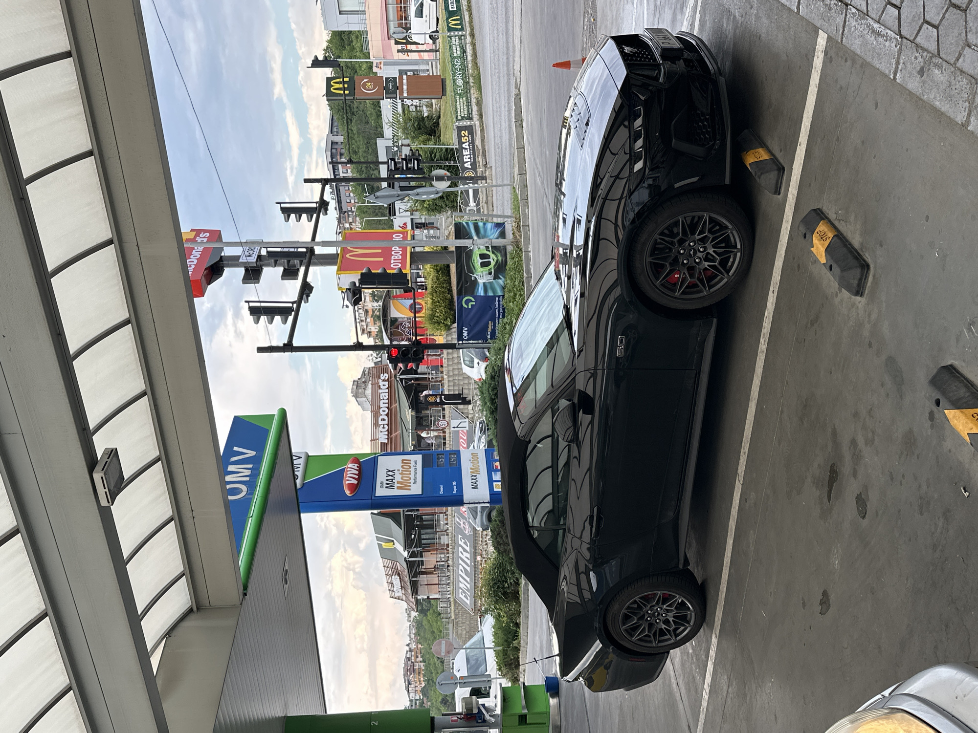 A black Ford Mustang convertible is parked at a gas station with McDonald's and other businesses visible in the background. The gas station appears to be an OMV, indicated by the signage. The Mustang is parked in a designated spot, with yellow chocks labeled "STOP" placed behind the rear wheels.
In the background, a McDonald's restaurant with its iconic golden arches is prominent, along with other signs for businesses like "dm" and "AREA52". The sky is partly cloudy, suggesting it is daytime. The overall scene is that of a typical urban or suburban environment with commercial establishments and transportation infrastructure. The image is oriented vertically, with the ground and parking lot at the bottom of the frame and the sky at the top. There are no people clearly visible in the image, and the focus is on the car and the surrounding environment. The text visible includes brand names of the businesses and information related to fuel prices at the gas station. The location is identified as Veliko Tarnovo, Bulgaria.