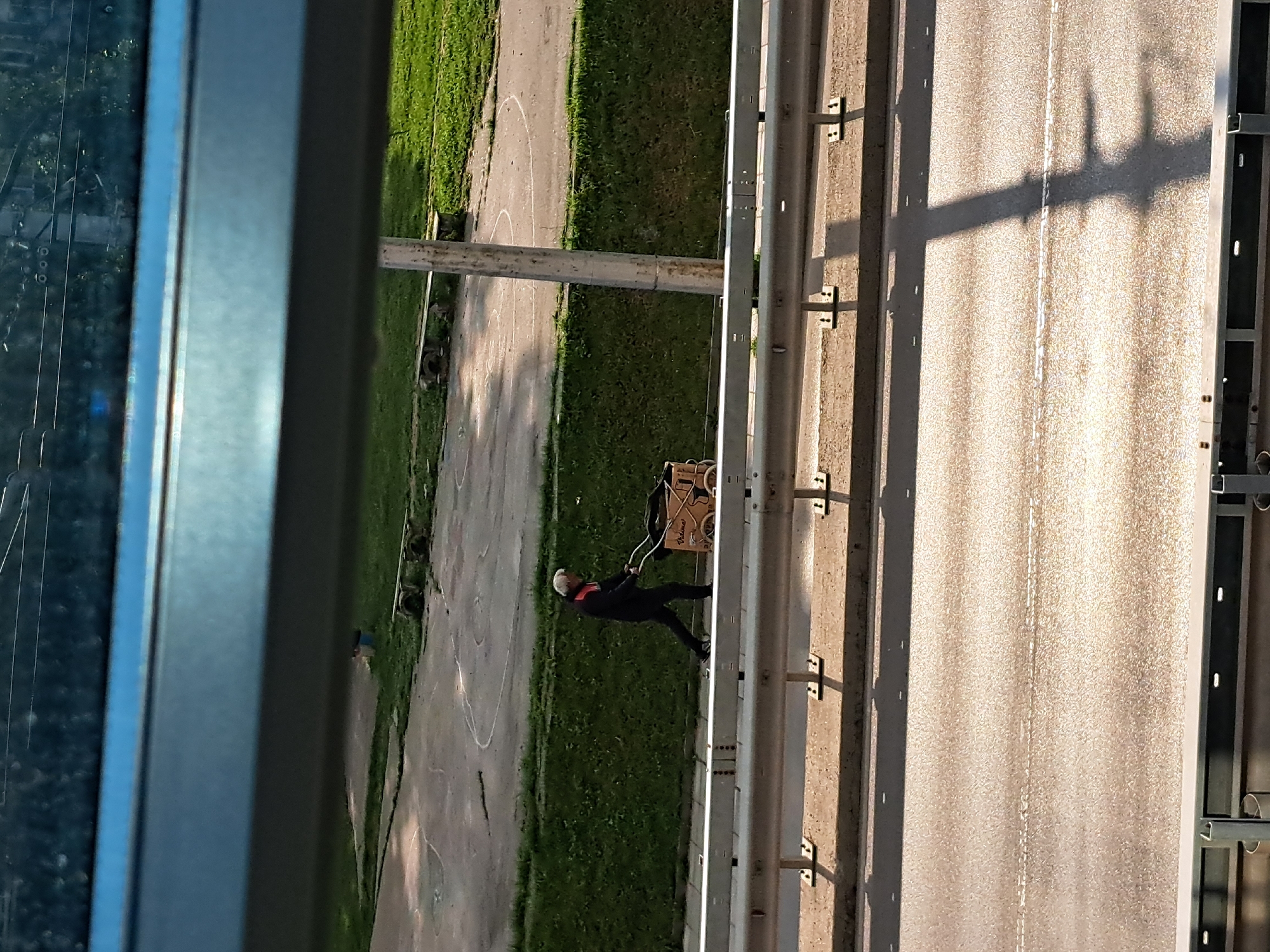 A low-angle shot shows a man pulling a cart on the side of a road, as seen through a window frame. The man, dressed in dark clothing with a white or gray cap, is pulling a cart with two wheels and what appears to be a wooden structure on top. He is walking along a road that runs alongside a grassy area with a circular paved section. A metal guardrail separates the road from the grassy area. The road surface has linear marks, possibly lane dividers. Shadows from a structure above fall across the road.