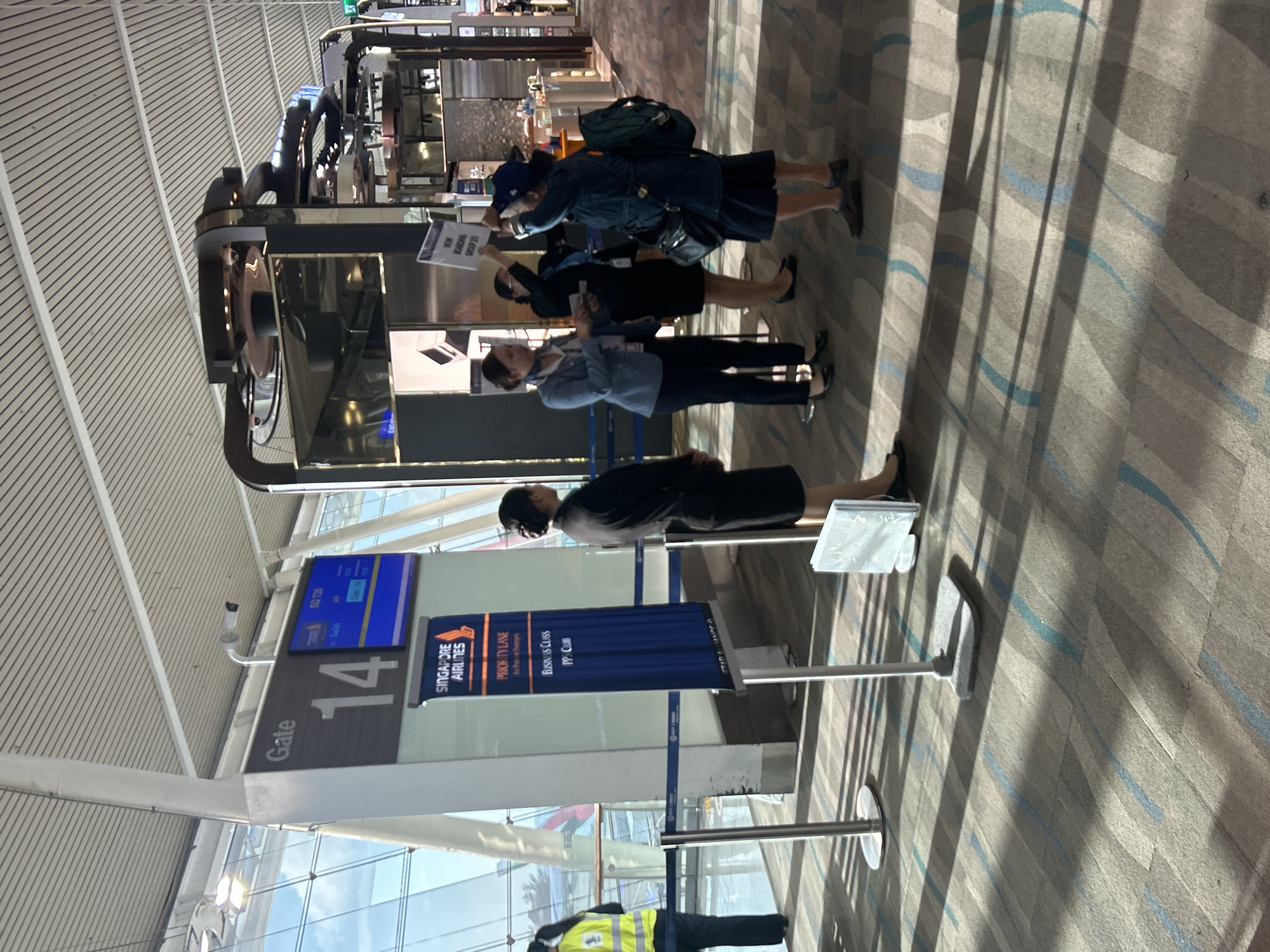 At an airport gate in Tambon Mai Khao, Thailand, a group of people are gathered. A large "Gate 14" sign stands prominently, with a Singapore Airlines banner below it indicating "Priority Lane," "Business Class," and "PPS Club." A flight information screen displays "SQ 735" and boarding details. Several individuals appear to be airline staff, some in uniform, managing the boarding process. One staff member is holding a sign that reads "NOW BOARDING GROUP 20." Passengers are seen in the background, and airport infrastructure, including ceiling panels and large glass windows, is visible. The floor is carpeted with a geometric pattern.