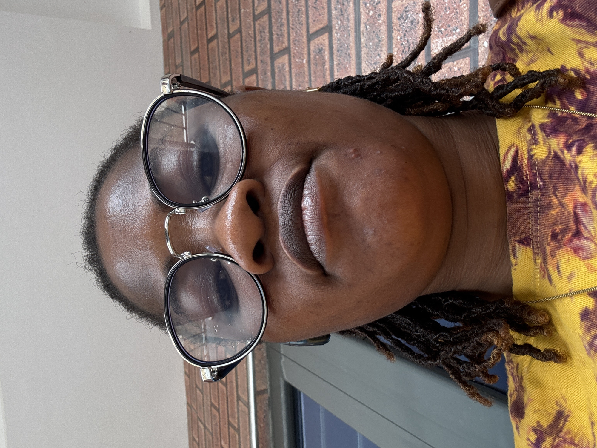 A low-angle, close-up selfie taken outdoors shows a Black person wearing glasses and a yellow t-shirt with a tie-dye pattern. The person has dreadlocks and gold hoop earrings. In the background, a brick wall is visible. The image was taken in Ajara, Nigeria.