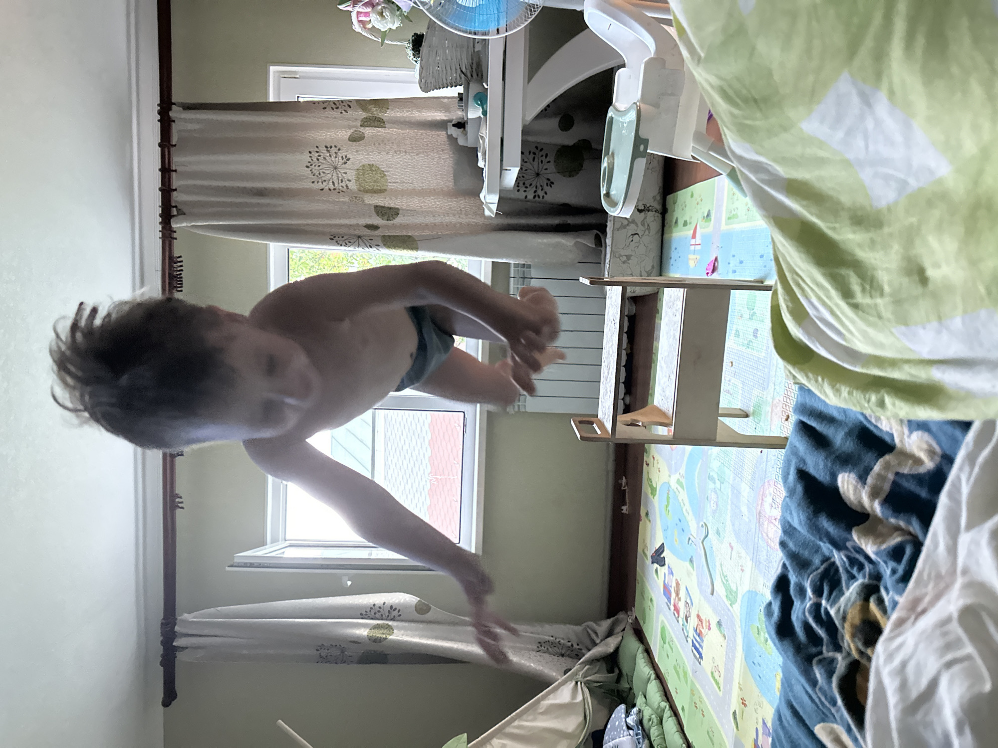 This media file captures a playful moment of a young child in what appears to be a bedroom or playroom. The child, seen from the torso up, is bare-chested and wearing dark shorts. Their hair is dark and somewhat disheveled, suggesting recent activity. The child's arms are extended, and their hands are blurred, indicating movement, possibly jumping or reaching.
The setting is a domestic interior. Behind the child, curtains with a subtle circular pattern are visible. A window with white frames allows natural light into the room. To the right of the child, a white high chair or similar piece of baby equipment is present, with a blue fan positioned above it. A playmat with a colorful road and cartoonish figures of children and animals is laid out on the floor. The playmat is partially covered by a light green and white patterned fabric, possibly a blanket or sheet. Further to the right, a dark blue fabric with an embroidered design is also visible.
The overall impression is one of a lively and active child engaged in play. The natural lighting suggests it might be daytime. The presence of the playmat and high chair points to a child-friendly environment, likely in a home. There are no visible text elements in the image. The rotation of the image suggests it was taken with a phone camera, possibly while the photographer was also in motion or the device was held at an unusual angle.