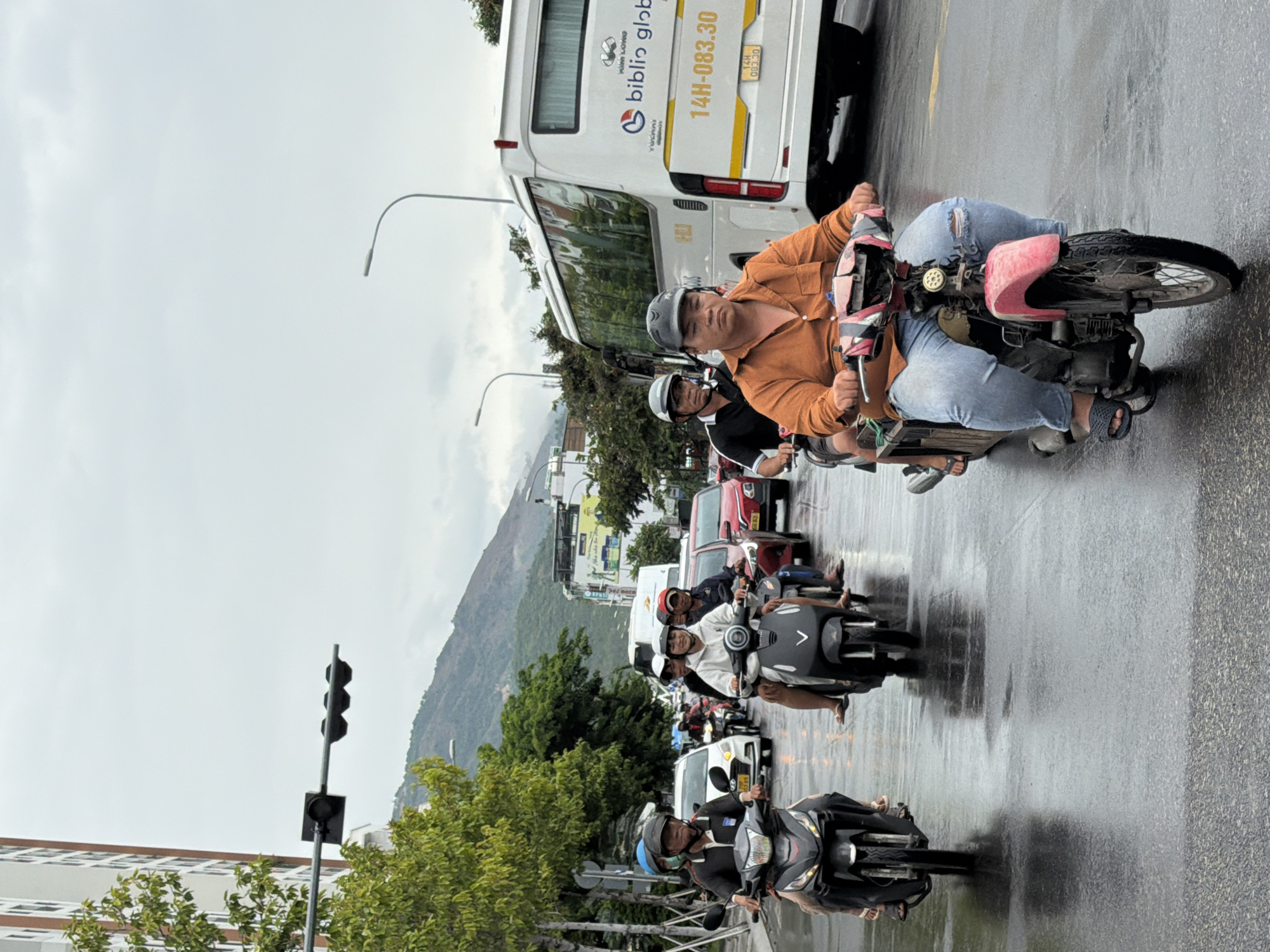 The image depicts a street scene in Nha Trang, Vietnam, characterized by overcast weather and wet asphalt roads, indicating recent rainfall. Multiple motorbikes are navigating the traffic, with riders wearing helmets and casual attire. In the foreground, a man in an orange shirt and light blue jeans rides a red and pink motorbike. Behind him, another man in a black and white shirt and helmet rides a dark gray motorbike. Further back, several other motorbikes and a few cars, including a white sedan and a red hatchback, are visible. A large white bus with "BIBLIO GLOBAL" branding and license plate "14H-083.30" is also present. Trees line the street, and a tall building with white and brown sections stands to the left. In the distance, mountains are shrouded in a light haze. Traffic lights are suspended over the intersection.