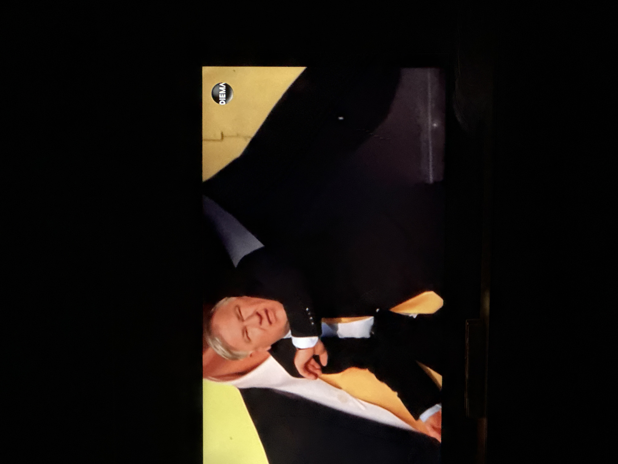 The image shows a light-skinned man in a black suit with white cuffs, lying on a golden surface. He appears to be on his side with his eyes partially closed. The lighting is dim, and a logo that reads "DIEMA" is visible in the upper left. The city of Sarnevo, Bulgaria is not visible in the image. The scene appears to be part of a broadcast or recording.