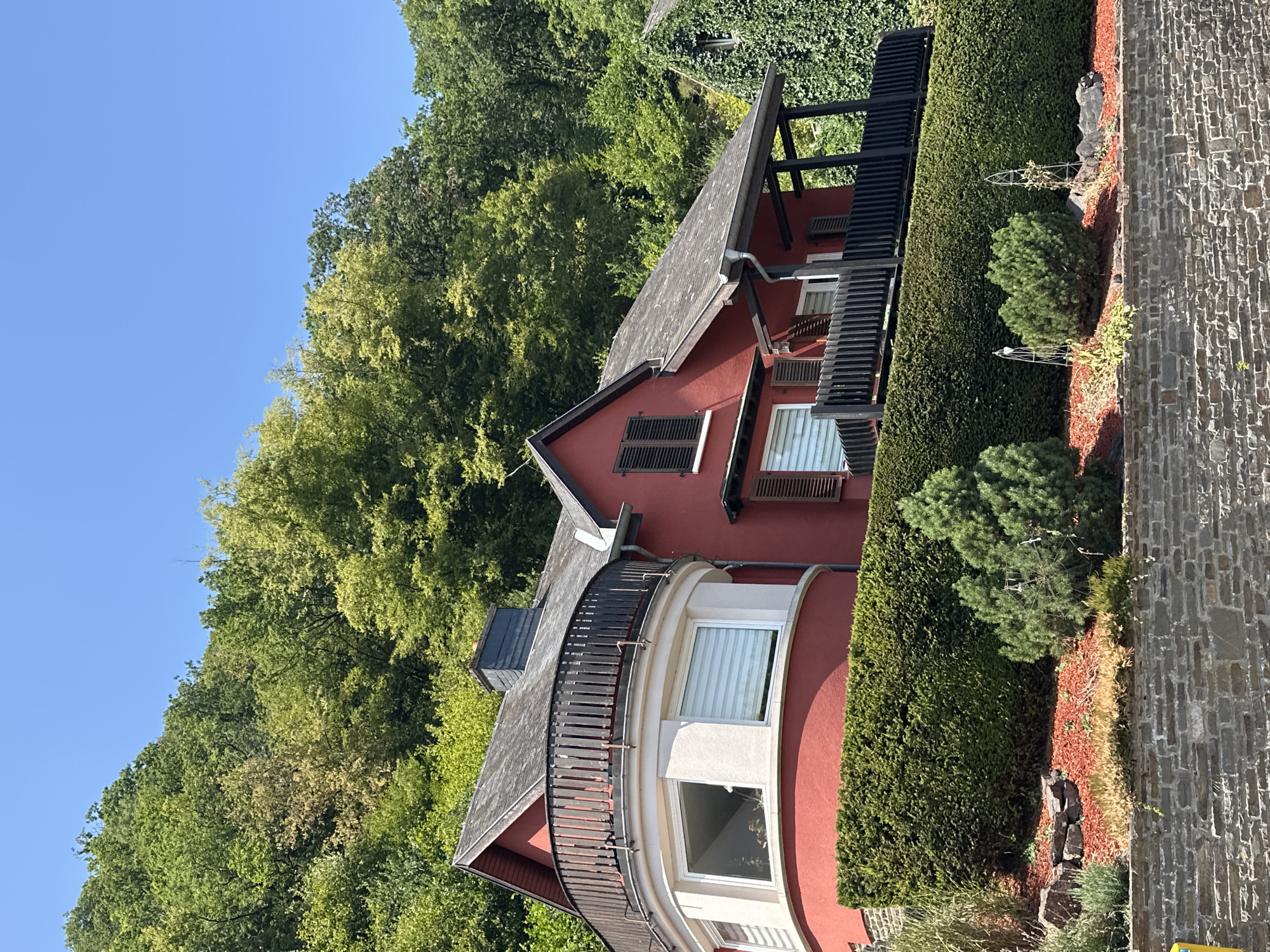 The image depicts a two-story red house situated on a hillside in Vianden, Luxembourg. The house features a dark, sloped roof and a prominent curved balcony with a dark wooden railing on the upper level. The lower part of the house has a large, curved bay window. A neatly trimmed green hedge runs along the front of the property, with red mulch and several small trees and shrubs in front of it. A stone retaining wall forms the base of the landscaped area. The house is surrounded by lush green trees that ascend the hillside behind it. The sky is clear and blue. There are no people visible in the image.