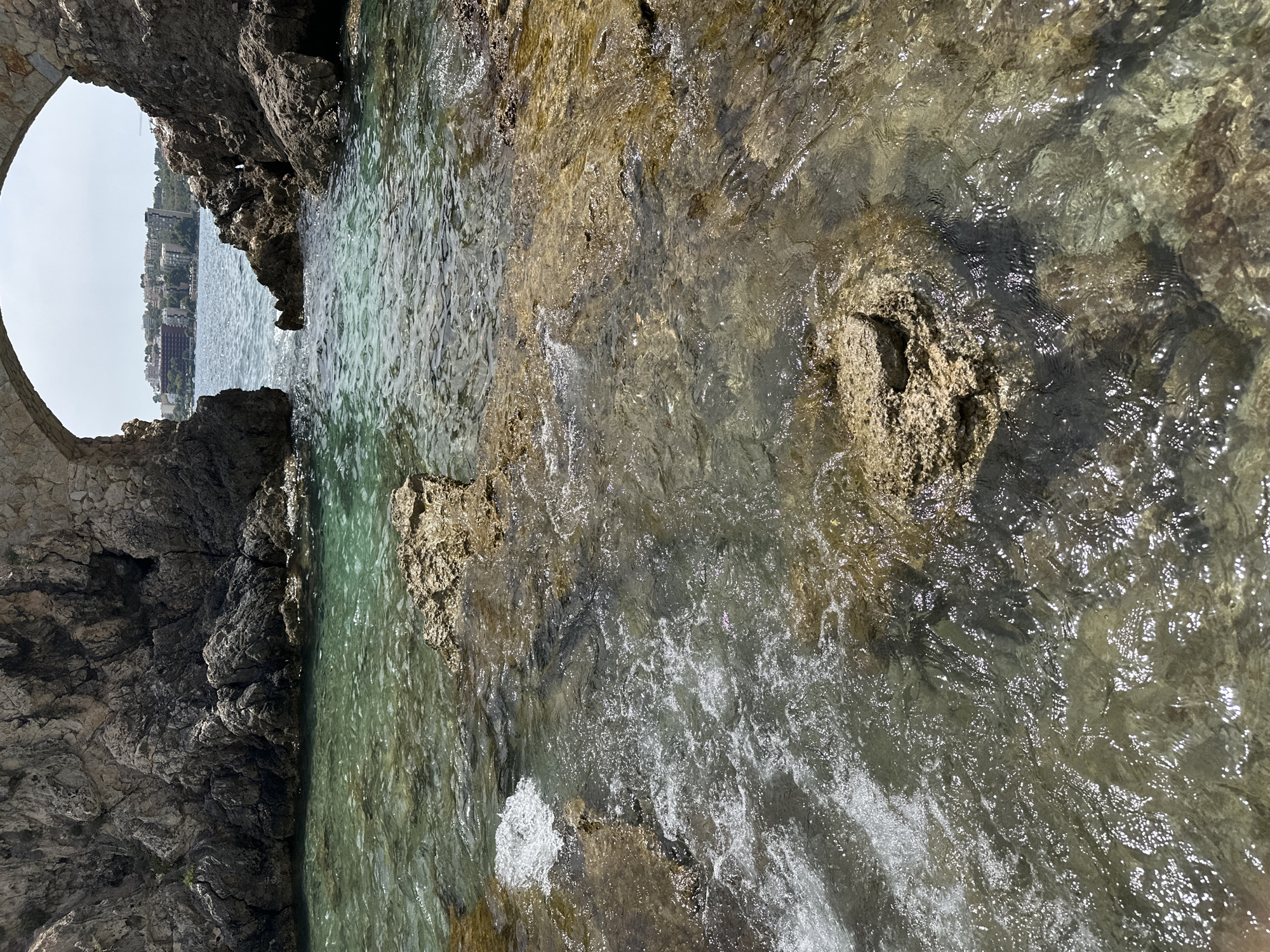 The image depicts a clear, shallow body of water with visible rocks beneath the surface, located in Sant Agustí, Spain. The water is a vibrant turquoise green near the rocky shore and becomes a deeper blue further out. Several large, dark grey rocks frame the foreground, with smaller, lighter brown rocks protruding from the water. The surface of the water is slightly rippled, with areas of white foam indicating movement, likely from gentle waves. In the background, seen through a natural rock arch, is a coastline with buildings and vegetation, suggesting a developed area. The sky is bright and appears to be a clear day. There are no people visible in the image.