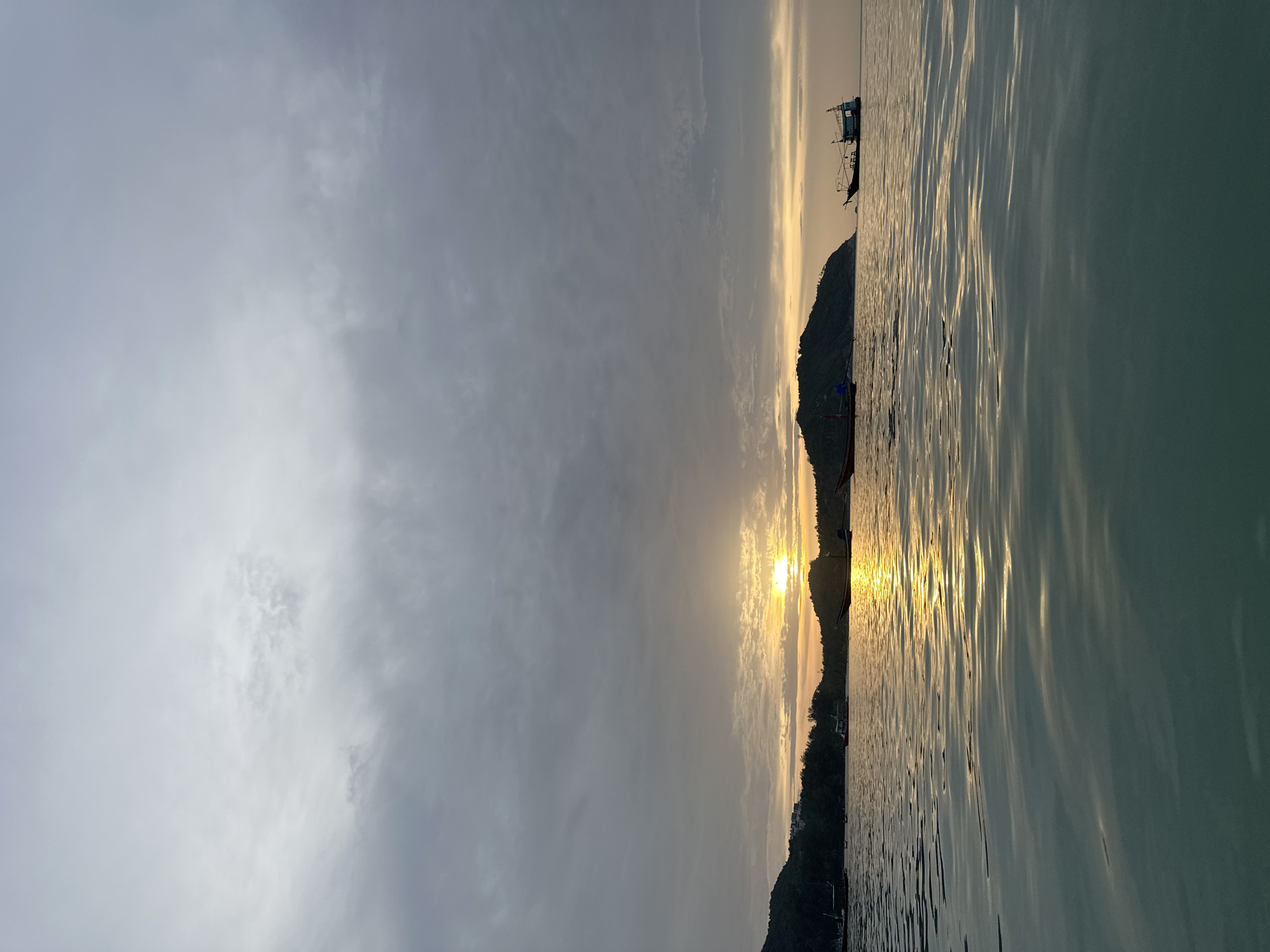 An image taken at sea off the coast of Koh Phangan District, Thailand, at sunrise. The sky is cloudy and gray, with the sun peeking through the clouds, casting a golden reflection on the water. The sea is calm, with gentle waves. In the distance, islands with lush green hills can be seen. Several boats are scattered across the water, some anchored and others moving slowly. One boat in the foreground appears to be a fishing vessel, while others are smaller, possibly longtail boats. The overall atmosphere is peaceful and serene, capturing the early morning light over the ocean.