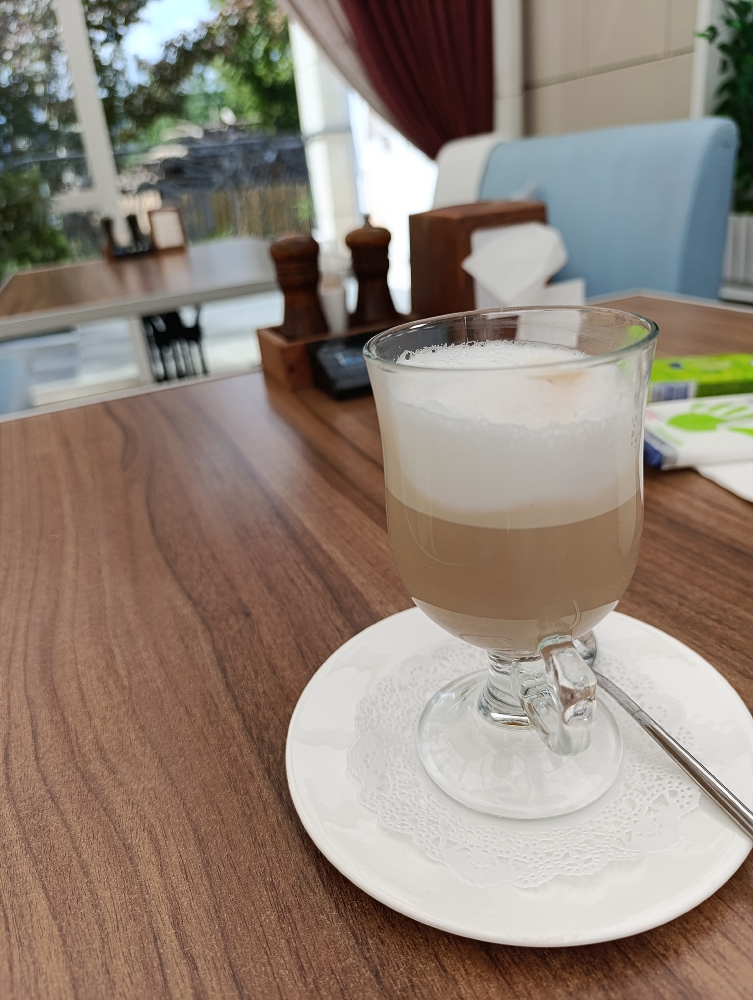 A tall glass of layered coffee drink, likely a latte or macchiato, sits on a white doily and saucer on a brown wooden table. The drink has distinct layers of coffee, milk, and foam. In the background, out of focus, are salt and pepper shakers, a tissue dispenser, and a blue upholstered chair. Through a large window, trees and sky are visible, suggesting an outdoor seating area or a cafe with a view. The overall setting appears to be a cafe in Chaikovsky, Russia. There are no people or active interactions visible in the image.