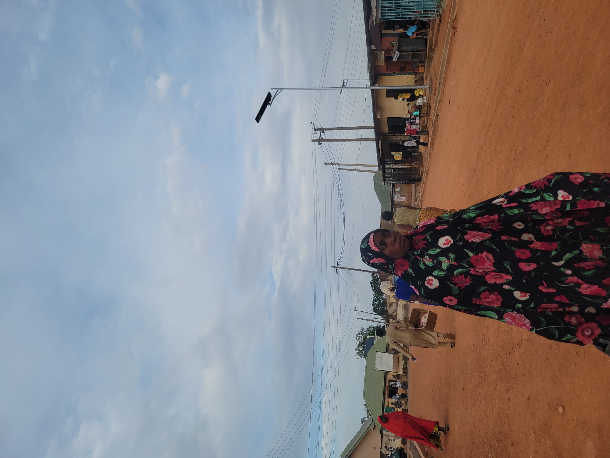 An outdoor, eye-level shot captures a vibrant street scene in Jos, Nigeria, featuring people, buildings, and infrastructure under an overcast sky. The image is taken at a slight angle, with the ground sloping gently upwards from left to right.
In the foreground, a woman wearing a black dress adorned with large pink and red floral patterns, and a matching headscarf, stands on a reddish-brown dirt road, looking directly towards the camera with a neutral expression.
The middle ground is dominated by a wide, unpaved reddish-brown road or path. Several other individuals are visible in the distance: one person in a light brown garment walks away from the camera, and another, wearing a striking red outfit, walks towards the right side of the frame. Along both sides of the road, various single and multi-story buildings line the street. These structures exhibit diverse facades and colors, with some having blue or green roofs, suggesting commercial or residential use. Several figures are discernible near these buildings, engaged in daily activities.
Overhead, a network of wooden utility poles with numerous power lines stretches across the sky, characteristic of local infrastructure. A modern, tall metal pole fitted with a solar-powered street light stands prominently on the left side, indicating the presence of public lighting. The sky is largely covered with light grey and white clouds, suggesting an overcast day with diffused daylight. No specific text is clearly legible on any signs or objects in the image.