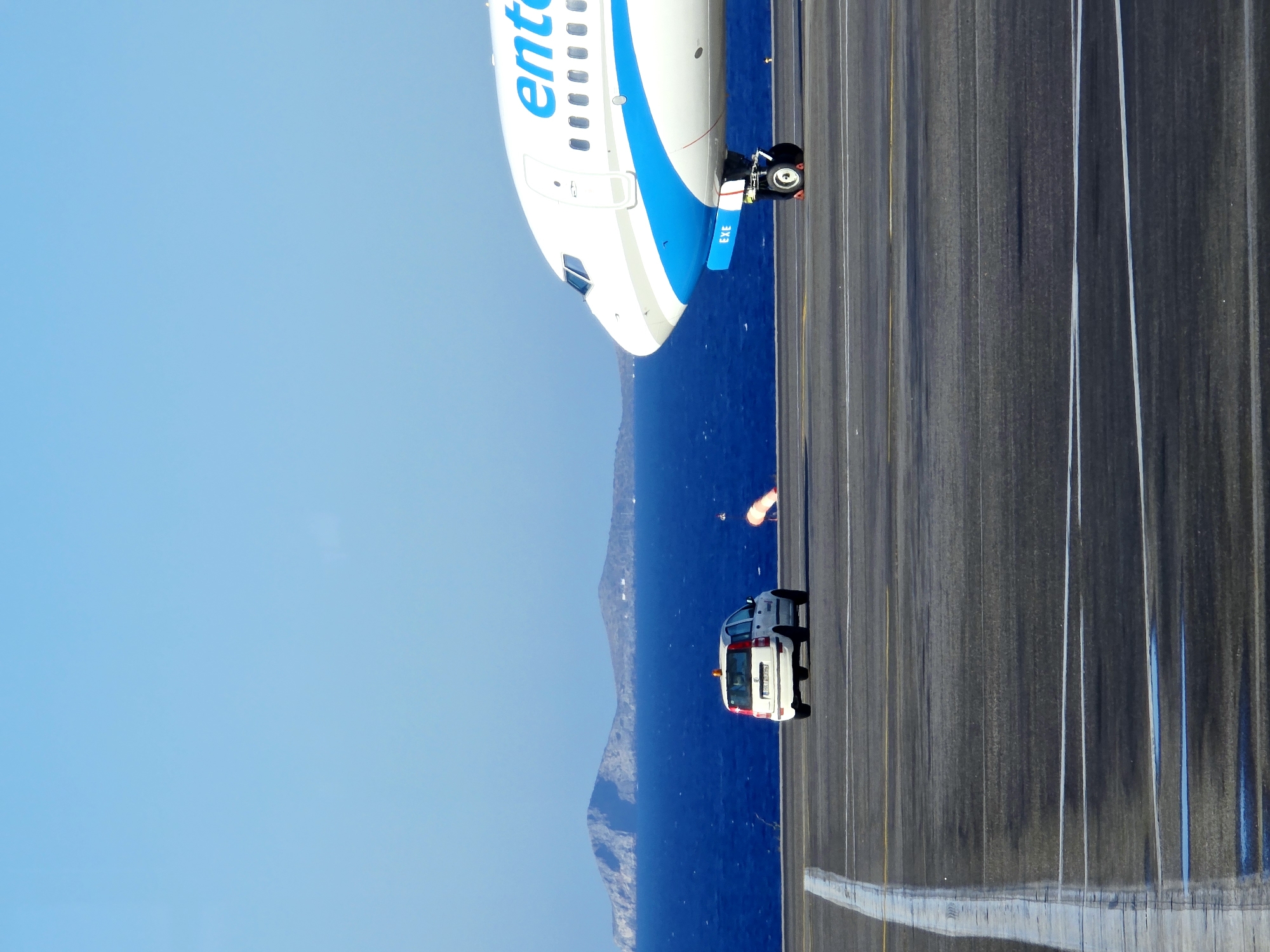An airplane is on a runway with the blue sea and islands in the background. A white car is also on the runway. The airplane is white with blue lettering that reads "ente." The car is white and has a light on top. The sky is clear and blue. The scene appears to be at an airport on a sunny day. The location of Nea Alikarnassos Municipal Unit, Greece is associated with Kos International Airport (KGS), which is situated on the island of Kos, in the Dodecanese archipelago. This airport serves the island of Kos and is the sixth busiest airport in Greece.