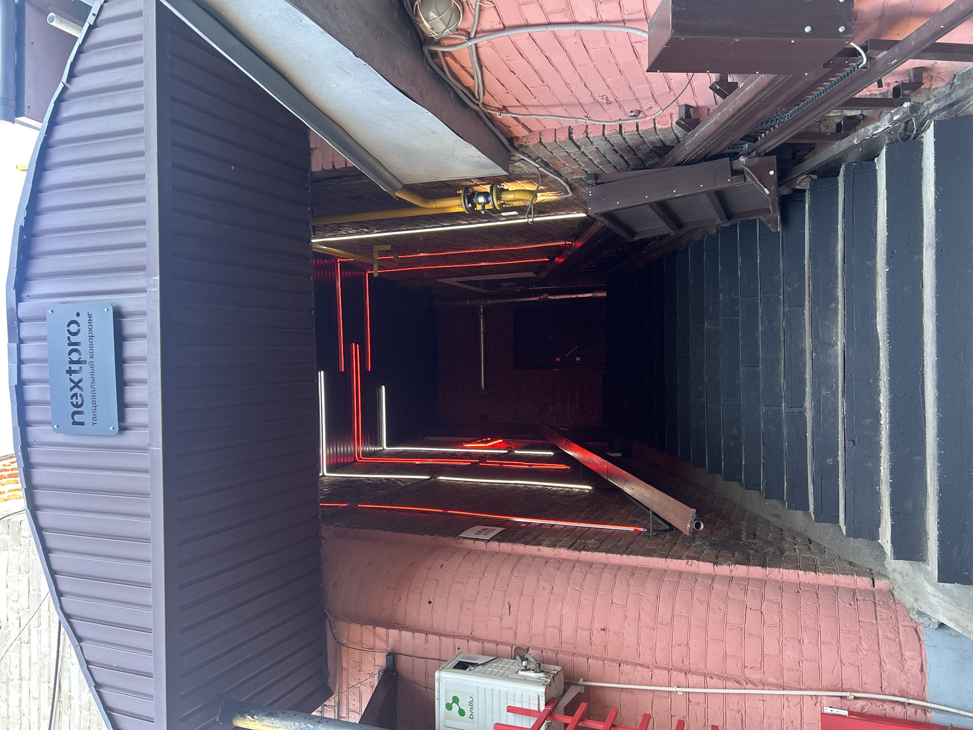 The image depicts an entryway in Izhevsk, Russia. To the left is a brown metal-sided structure bearing a sign with the text "nextpro." and some smaller Cyrillic script. The entryway leads down a short flight of black-painted steps alongside a pink-painted brick wall. The entry appears to be into a basement or lower-level establishment, indicated by the neon lighting within: alternating bands of red and white are installed on the floor. Overhead, the space is crowded with infrastructure like wiring, ductwork, and what appears to be a metal beam. Air conditioning units are placed along the pink brick exterior wall.
