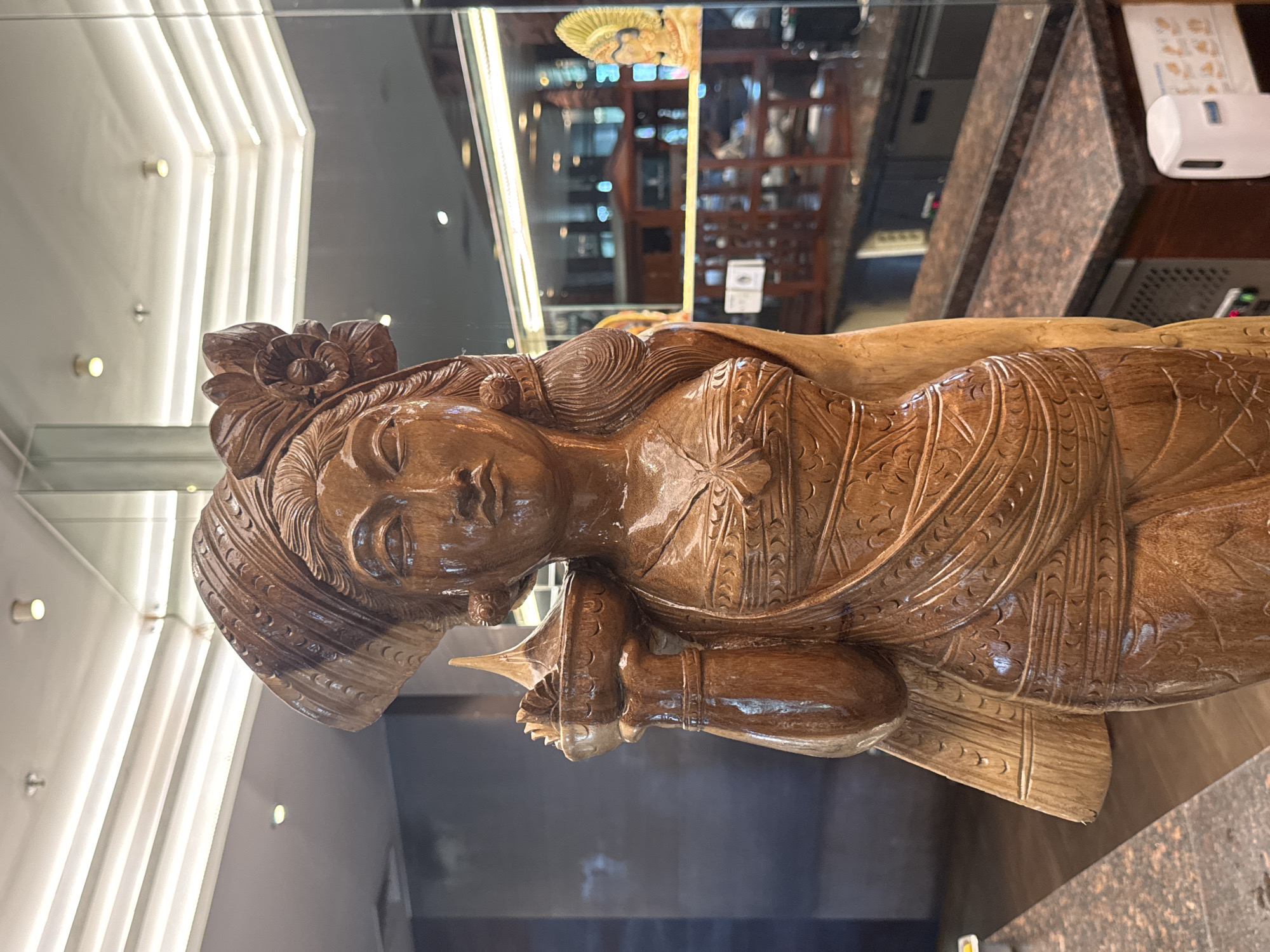 A wooden statue of a woman wearing a traditional headpiece and holding a bowl is displayed in front of a blurred background. The statue is carved with intricate details on her clothing and jewelry. The background appears to be an indoor setting with lights on the ceiling and a reflection of what looks like a restaurant or shop. In the reflection, another smaller statue or artwork is visible, as well as kitchen counters and equipment. The lighting is bright, casting a warm glow on the wooden statue.