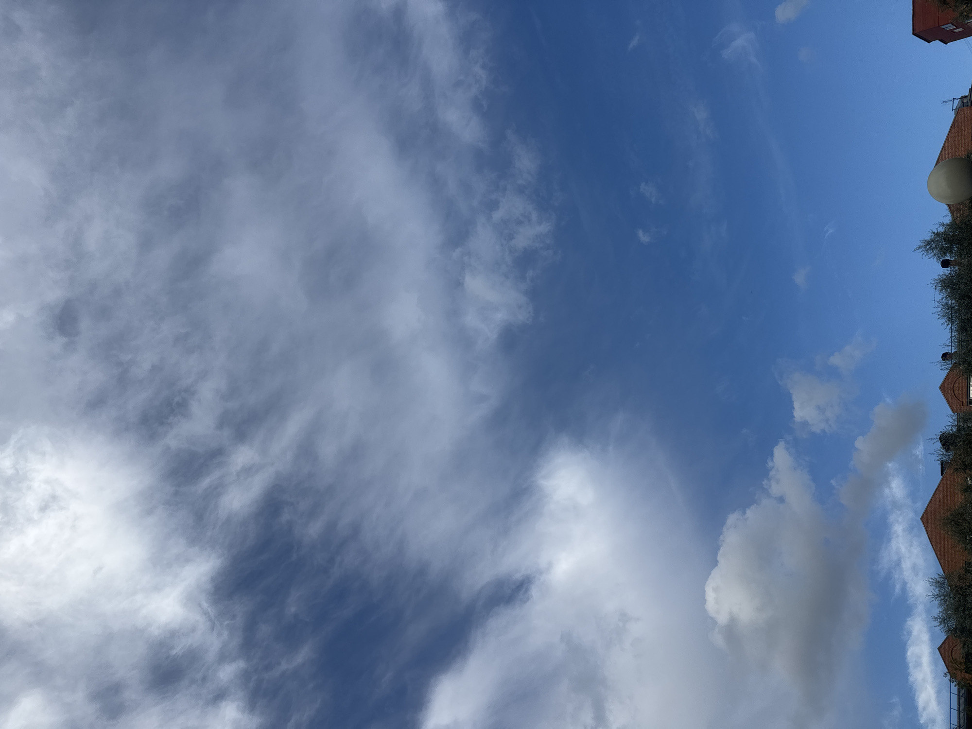 This image captures a view of the sky above Sovkhoza imeni Lenina, Russia. The sky is predominantly blue with a mix of clouds. Wispy cirrus clouds stretch across the upper portion of the frame, interspersed with denser, more opaque clouds in the lower half. In the foreground, the rooftops of several buildings are visible, some with trees and foliage in front of them. The architecture suggests a residential or possibly commercial area. The overall scene depicts an outdoor environment under a dynamic sky.