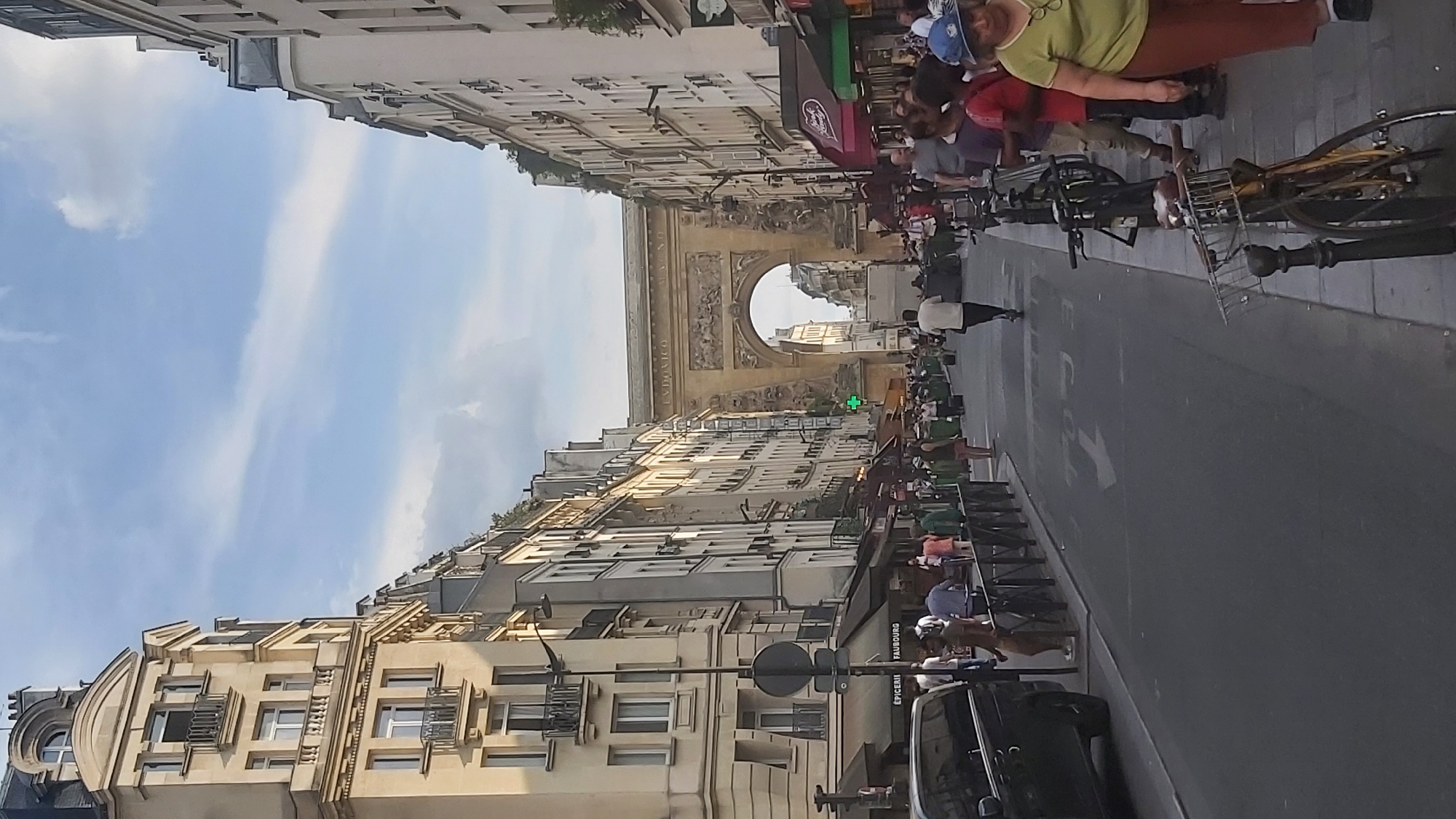 The image shows a street scene in Paris, France, with the Arc de Triomphe du Carrousel visible in the distance. On the left, a light-colored building with multiple floors and balconies lines the street. On the right, similar buildings with shops and awnings are present. A black van is parked on the left side of the street. Numerous people are walking along the sidewalk and crossing the street. Several bicycles are parked on the right side of the road. The street is paved with asphalt and has a white arrow painted on it. A green cross symbol is visible on the right side of the street, indicating a pharmacy. The sky is partly cloudy.