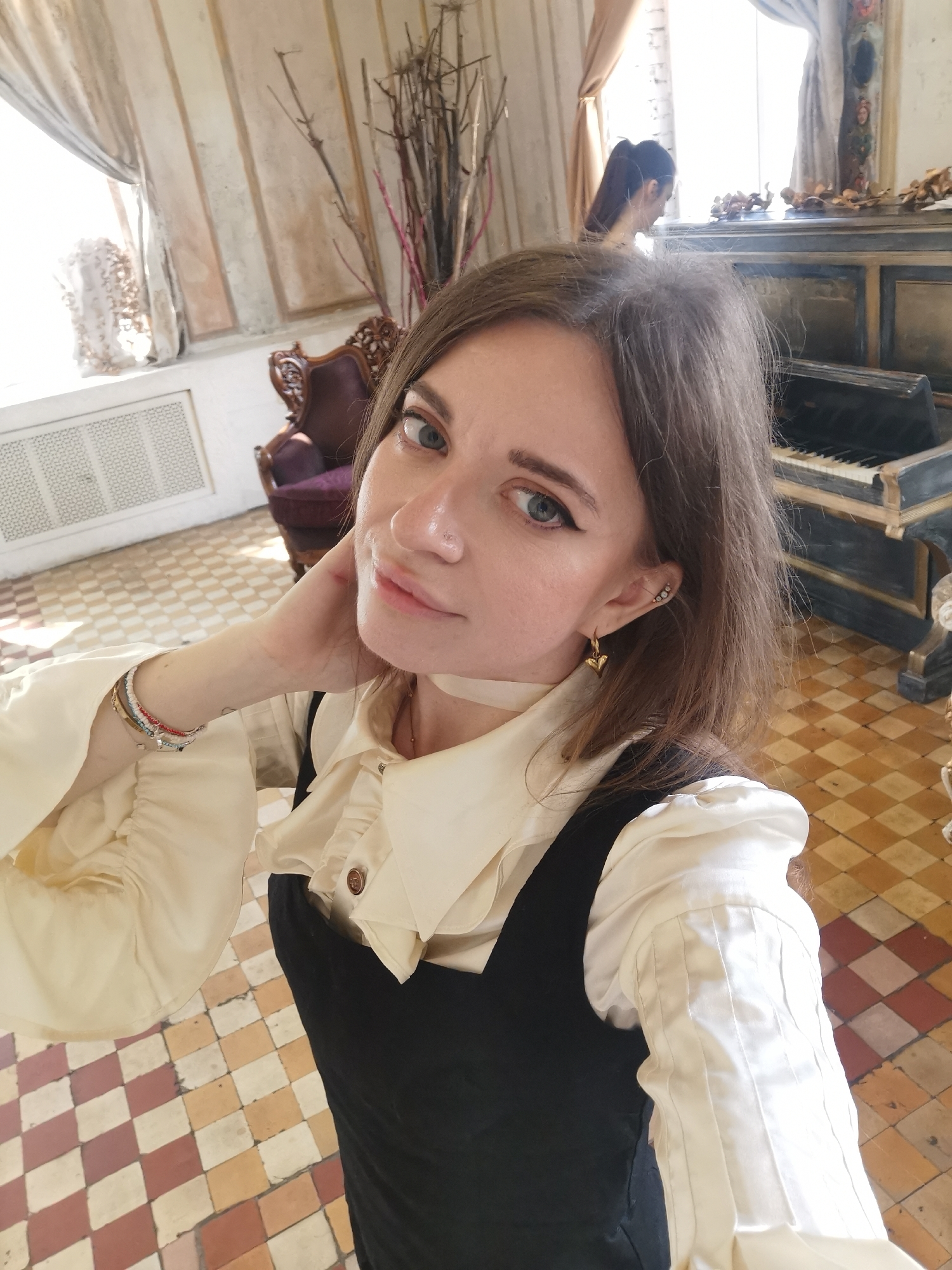 A woman with brown hair and blue eyes is pictured indoors, posing for a selfie. She is wearing a cream-colored blouse with ruffled sleeves and a dark pinafore dress. She is adorned with jewelry including a choker, earrings, and bracelets on her left wrist. She has her left hand raised to her neck.
Behind her, a tiled floor can be seen. There's a vintage-style wooden piano, a decorative arrangement of branches, and an antique chair with a purple cushion. In the background, a person is visible near the piano. The room has patterned wallpaper and curtains.
Given the architectural style and decor, this image appears to be taken in Moscow, Russia. The presence of the vintage-style piano, chair, and patterned walls suggest the photo may have been taken in an older building such as a museum or a historical place in Moscow.