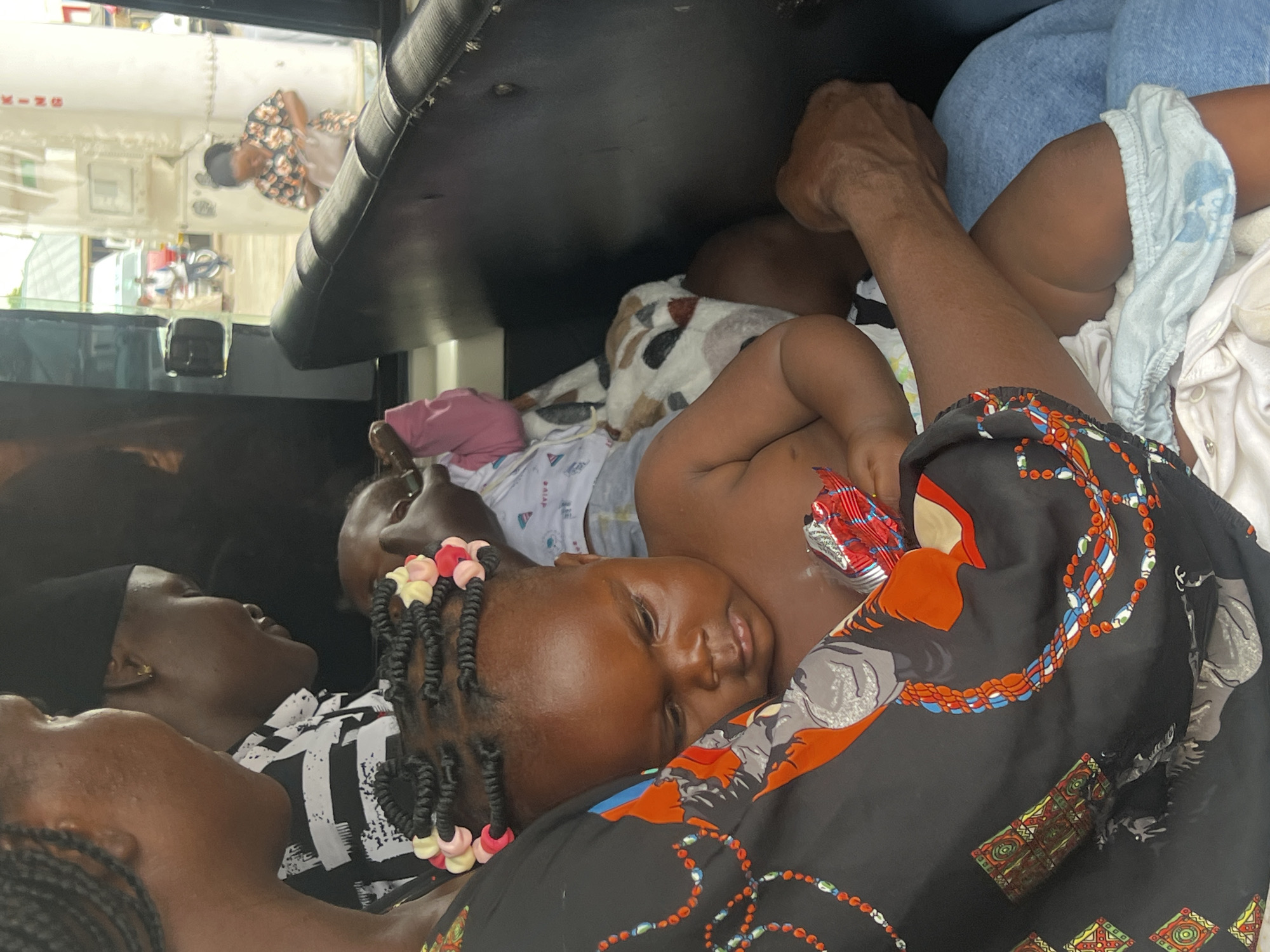 The image shows a group of people, likely a family, in what appears to be a cramped indoor space, possibly a vehicle. The lighting suggests it could be daytime.
The primary focus is on a young child lying on their side, nestled among several adults. The child, with dark skin and braided hair adorned with colorful beads, is resting with their eyes closed, appearing peaceful or asleep. Another child, also with dark skin, is visible behind the first, partially obscured. Two adults are also present, their faces close to the children. One adult, wearing a black headscarf, is looking away from the camera, while another adult's arm is visible in the frame. The adults are dressed in various clothing, including a black and white striped shirt and a black garment with a colorful, patterned design featuring chains and figures.
The setting seems to be confined, with the people positioned close together. The background shows parts of what could be the interior of a vehicle, with a dark ceiling and a reflection in a window. A person wearing a patterned shirt is visible in the reflection. Objects like blankets or cloth are spread around the people, contributing to the crowded atmosphere.
The scene conveys a sense of intimacy and closeness, possibly indicating a journey or a moment of rest. The expressions on the visible faces are relaxed, suggesting a calm and familial environment. No visible text is present in the image.
