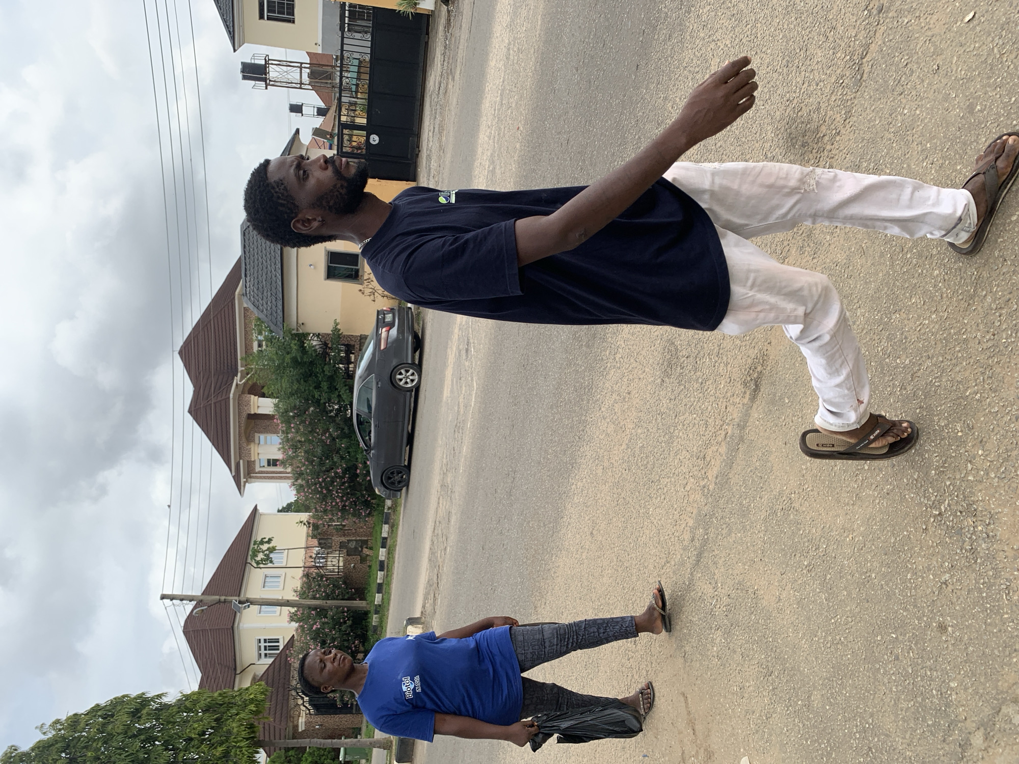 In Abuja, Nigeria, a man and a woman are walking on a paved road under a cloudy sky. The woman, on the left, wears a blue t-shirt and patterned leggings, carrying a black plastic bag. She has dark hair pulled back and is looking downwards. The man, on the right, is in a dark t-shirt and light-colored pants. He has dark, short hair and a beard and is looking forward, walking ahead of the woman. In the background, there are modern houses with brown roofs, lush green trees, and parked cars. The environment appears to be a residential area within the city.