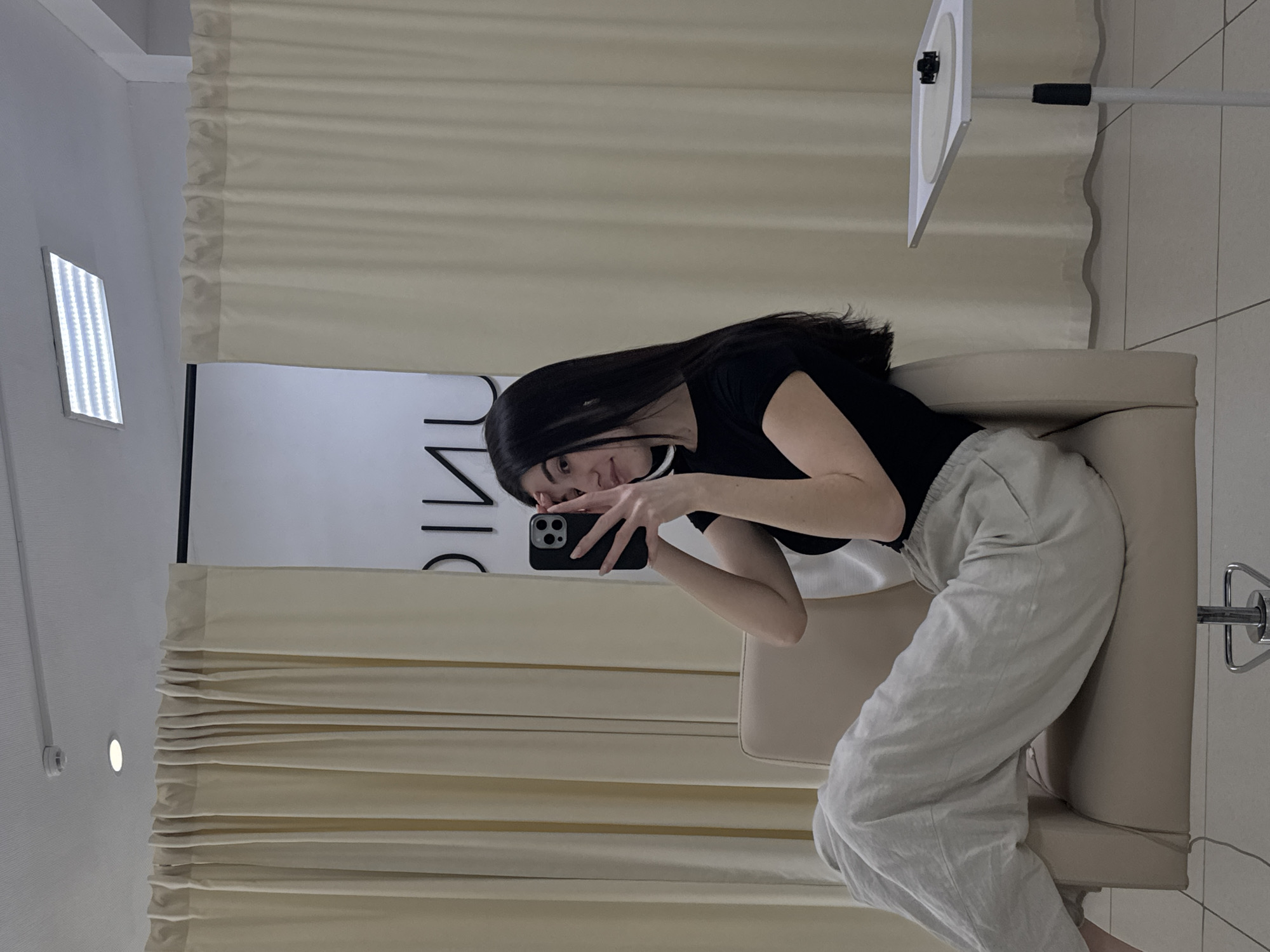 In the city of Chaikovsky, Russia, an indoor scene depicts a female subject taking a mirror selfie. The subject, positioned centrally, is a young adult female with long, straight dark hair, wearing a black short-sleeved top and light grey, loose-fitting trousers. She is seated in a light beige, upholstered salon-style chair with a chrome base, extending her legs towards the left foreground. She holds a black-cased smartphone with a triple-lens camera array in both hands, partially obscuring her face as she angles it for the reflection. Her visible right eye is directed towards the camera.
The background features light beige, pleated curtains hanging from a dark rod, dividing the space. Behind the curtains on the right, a white wall is visible, displaying inverted black letters that appear to spell "ƆINU", suggesting a reflection of the word "UNIC" or similar. The ceiling is white, equipped with a small, round recessed light, a rectangular LED panel light, and a white, cylindrical sensor mounted on a track. The floor consists of light-colored square tiles with dark grout lines. To the subject's right, a white, adjustable-height stand with a square top holds a white circular disc and a small black object.