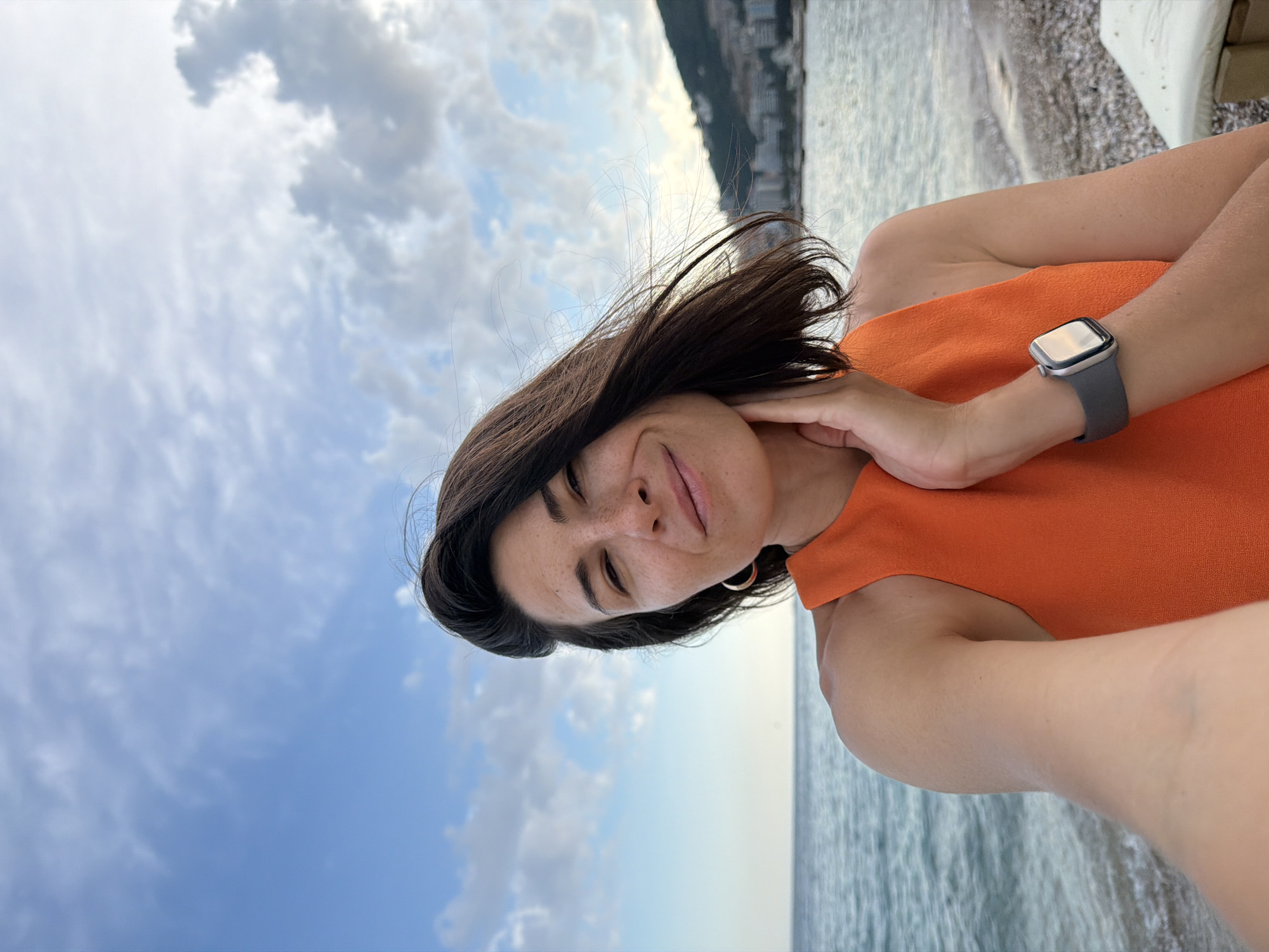 A woman with dark hair and fair skin, wearing an orange sleeveless top and a grey smartwatch, is taking a selfie on a beach. She is standing with the sea and a coastal city in the background. The sky is cloudy. The city in the background is Budva, Montenegro.