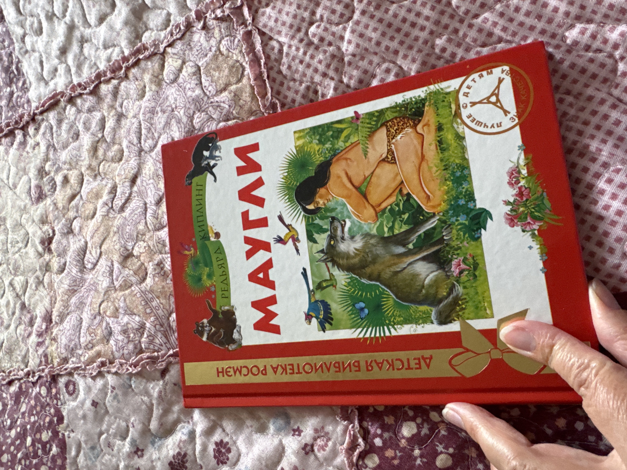 A red book titled "МАУГЛИ" (Mowgli) is displayed on a quilted pink and white blanket. The book cover features an illustration of a young man with dark hair, wearing a leopard print loincloth, crouching in a jungle setting. He is looking at a wolf. Several birds and butterflies are also depicted in the scene. The spine of the book has text in Russian that reads "ДЕТСКАЯ БИБЛИОТЕКА РОСМЭН" (Children's Library Rosmen). A circular seal with Russian text is visible on the bottom right of the cover. A person's hand is visible on the left side of the image, touching the book. The environment appears to be indoors, with the book laid on a textured surface. There is no information in the image to suggest the location is the city of Ufa, Russia.