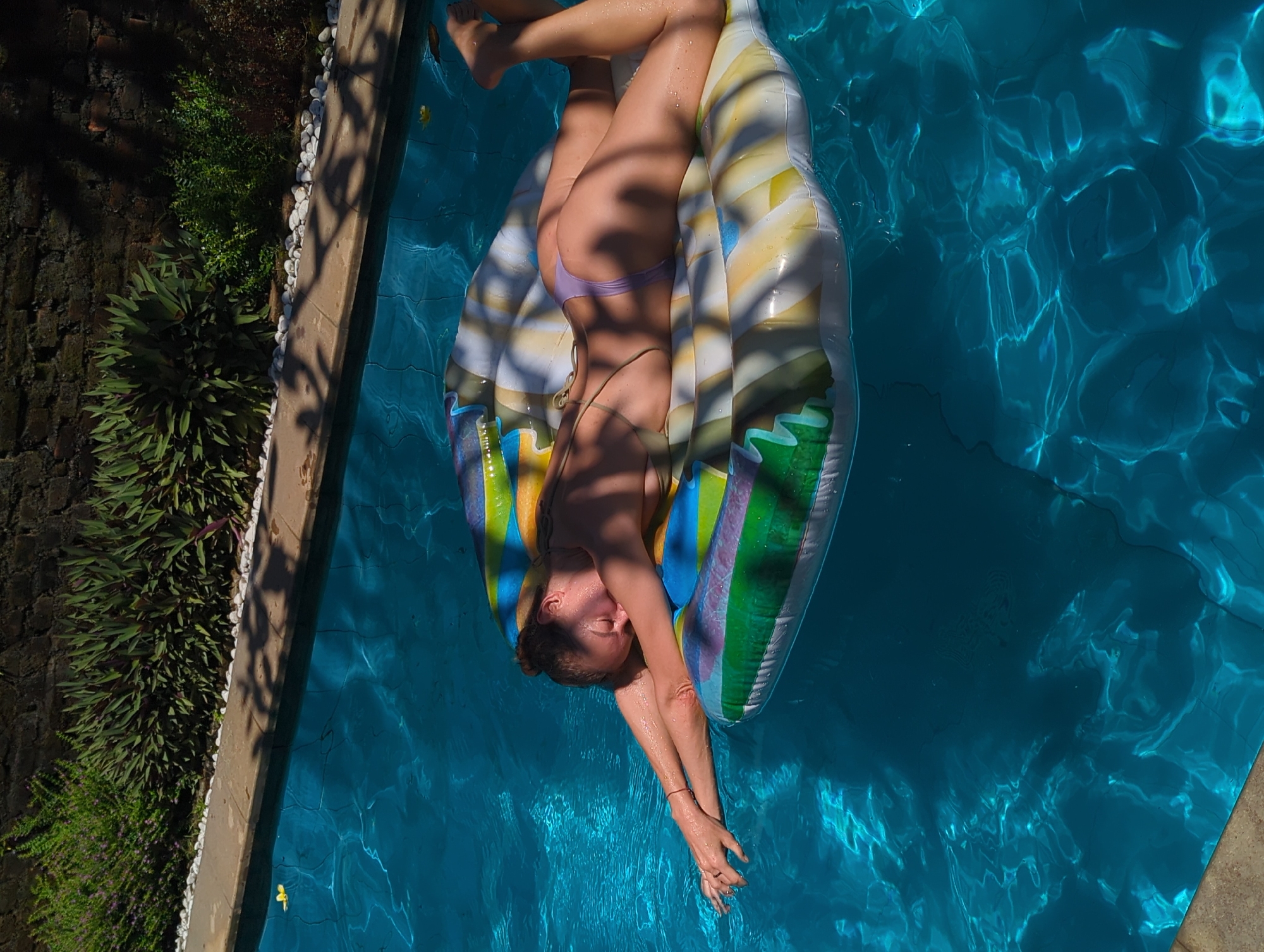 A woman reclines on an inflatable raft in a swimming pool. She is wearing a bikini and is oriented lengthwise on the raft with her head toward the bottom of the frame and her feet toward the top. Her arms are outstretched, and her hands are near the pool's surface. The raft is multicolored and reflects the sunlight. The pool water is blue and shows reflected light patterns. Along the left edge of the frame is a stone wall with plants and vegetation growing against it. The pool is located in North Kuta, Indonesia.