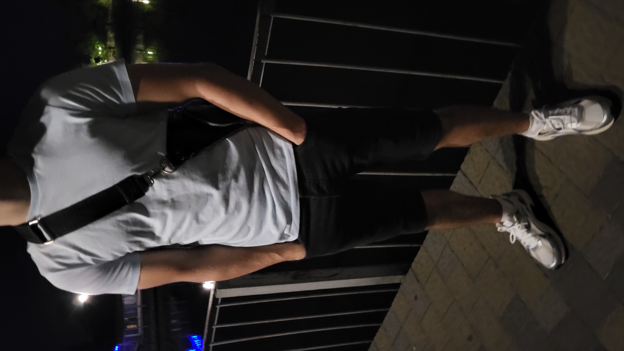 A person, whose face is not visible, stands on a paved surface next to a metal railing. They are wearing a light blue t-shirt, black shorts, and white sneakers with silver accents. A black crossbody bag with a silver buckle is slung across their chest. The background shows a body of water, possibly a lake or canal, with distant lights and trees, suggesting an outdoor urban setting at night. The lighting is dim, with artificial lights casting reflections on the water.