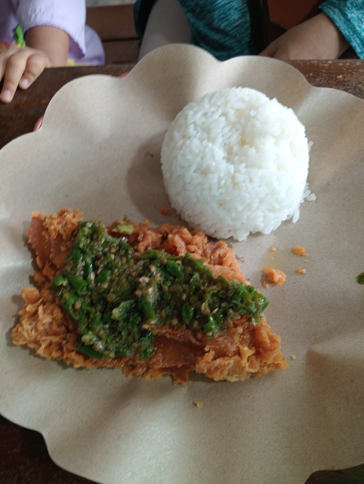 In Campago, Indonesia, a close-up shot shows a meal of fried chicken and rice served on a brown paper plate. The fried chicken, coated in a crispy batter, is topped with a vibrant green chili sauce. A mound of fluffy white rice sits to the right of the chicken. In the background, out of focus, are the arms of two individuals. One arm, on the left, is adorned with a light purple sleeve, while the other arm, on the right, wears a teal-colored garment. The meal is presented on a wooden table, suggesting a casual dining setting.