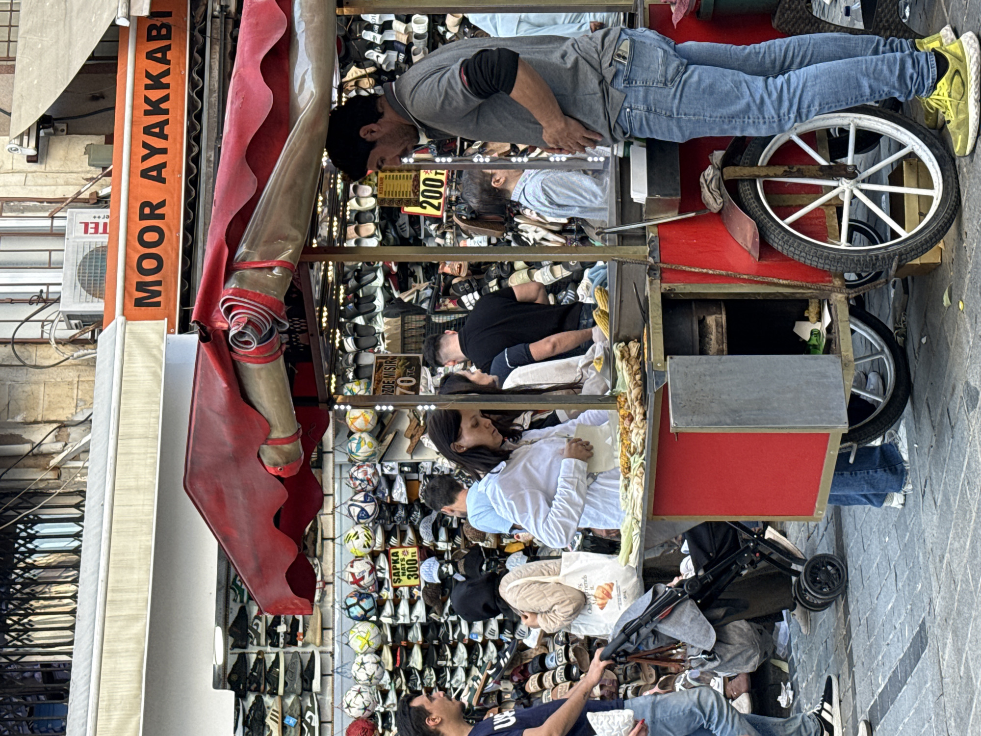 Here is a detailed description of the image:
**Scene:** The image shows a bustling street scene, likely a commercial area, based on the various street vendors. The foreground is dominated by a vendor's cart laden with items, mainly shoes and soccer balls. People are milling around the cart, either inspecting the merchandise or passing by.
**Vendor's Cart:** The cart is red with a white-spoked wheel. Shelves contain footwear in various styles. At the top are soccer balls. Signage is visible that prices items (e.g., "SAPKA HATS 300"). The front area appears to be selling grilled corn on the cob.
**People:** There are several individuals in the frame. Some seem to be customers browsing the goods on the cart. Others are simply passers-by. The individuals are of varying ages and are dressed in everyday casual clothing. One man is bent over, seemingly inspecting the shoes.
**Environment:** The background is a mix of building facades, suggesting a commercial area. There is a building with a sign "MOOR AYAKKABI." The street pavement is visible as well.
**General impression:** The scene is active, colorful, and gives the impression of a vibrant commercial area.
**Location Clues:** Based on the signs being partially in Turkish (e.g., "AYAKKABI," "SAPKA," "MISIR"), it's highly likely this is a street scene in Turkey. "MOOR AYAKKABI" might be the name of a shoe store in the vicinity. Unknown and Unknown information is unable to be discerned from the image.