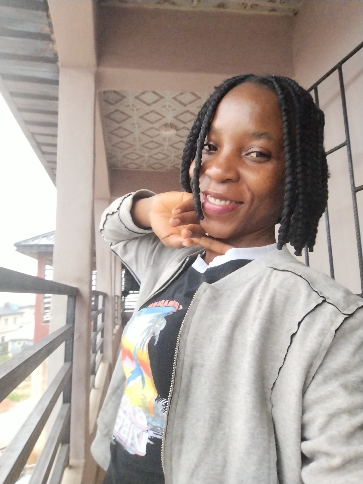 A medium shot photograph captures a young Black woman standing on a balcony, her body angled towards the camera. She has dark skin, braided hair, and is smiling broadly with her teeth showing. Her right hand is raised, touching her cheek and chin area. She is wearing a gray zip-up jacket over a black t-shirt with a colorful graphic print. The balcony railing is visible in the foreground, and a building with a patterned ceiling is in the background. The overall lighting is bright, suggesting it is daytime. The scene appears to be in Uselu, Nigeria.