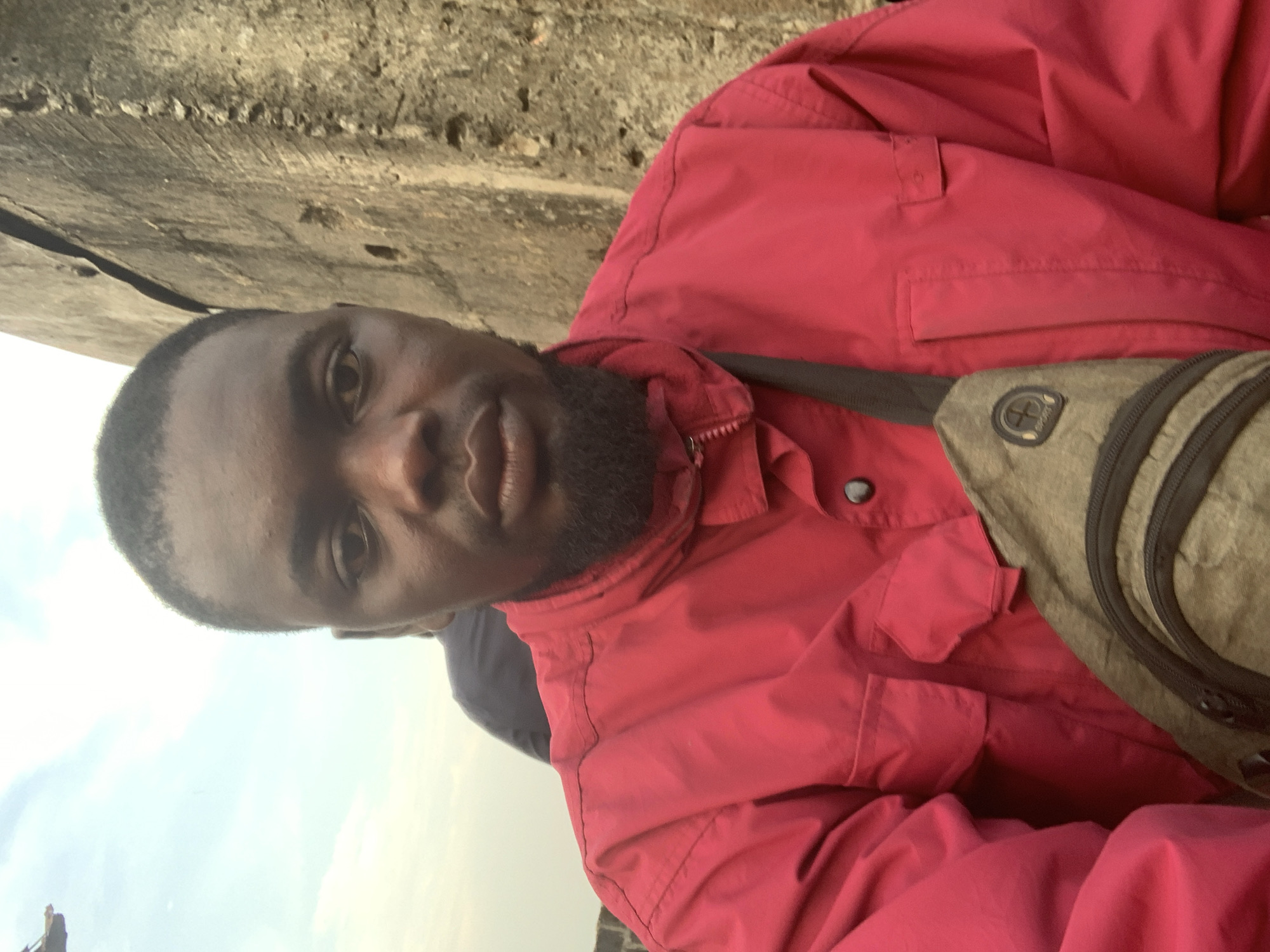 A selfie taken from a low angle shows a Black man from the chest up. He has short black hair and a beard, and is wearing a bright red jacket and a tan crossbody bag. The background shows a beige concrete wall and a cloudy sky. Another person's shoulder is visible behind the man's right shoulder. The photo was taken in Shagamu, Nigeria.