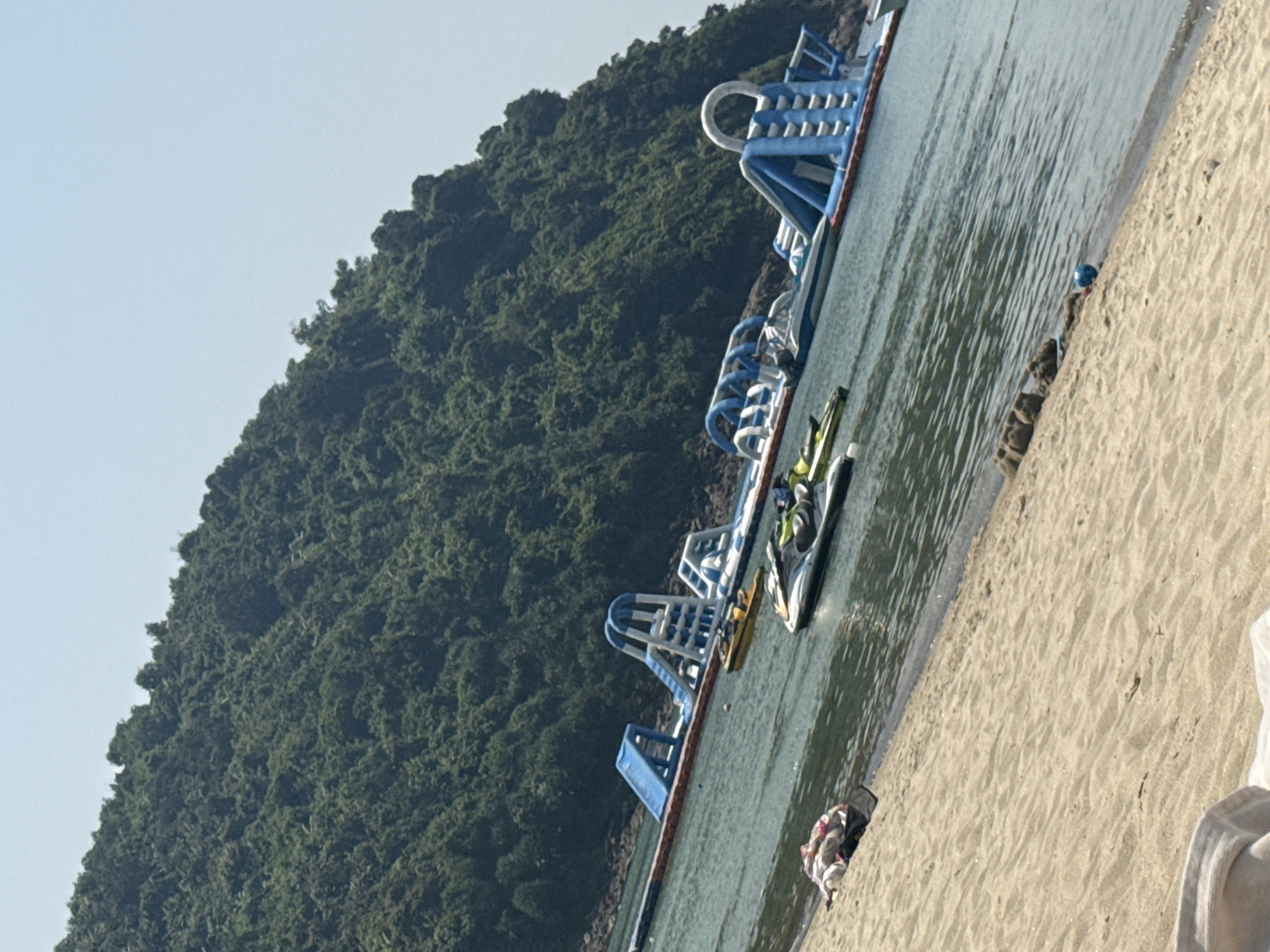 The image displays a sunny coastal scene in Đà Nẵng, Vietnam, specifically identifiable as part of the Sơn Trà Peninsula area due to the distinctive heavily forested mountain meeting the sea. The foreground features a light tan sandy beach sloping gently towards calm, clear greenish-blue water. On the sand, in the lower left, a pile of multi-colored fabric is visible. Towards the lower right, near the waterline, a small blue ball rests beside some amorphous sand structures.
In the midground, floating on the water, is a large, multi-sectioned inflatable water park structure, predominantly blue and white, consisting of slides, climbing ramps, and arched pathways. To the left of this structure, closer to the shore, a white and black personal watercraft (jet ski) with green accents is partially submerged. A yellow kayak or small boat floats further out, positioned between the jet ski and the inflatable park. A third, smaller, dark-colored watercraft is visible further offshore near the inflatable park.
The background is dominated by a steep, heavily vegetated mountain, densely covered in dark green foliage, rising directly from the water's edge and extending upwards to meet a clear, light blue sky. No individuals are visible within the frame.