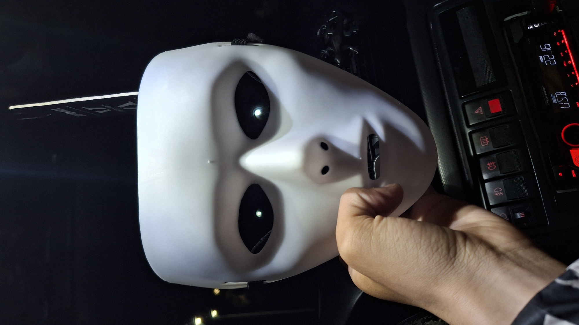 A stark white, featureless mask is being held by a light-skinned hand with a zebra-print cuff visible. The mask has large, dark eye sockets and small, nostril-like holes. The scene appears to be inside a vehicle, with a car's dashboard and console to the right, featuring illuminated buttons and a Pioneer radio display showing "22:16" and "USB." The interior is generally dark. There is no indication in the image that would connect it to the city of Shumen, Bulgaria.