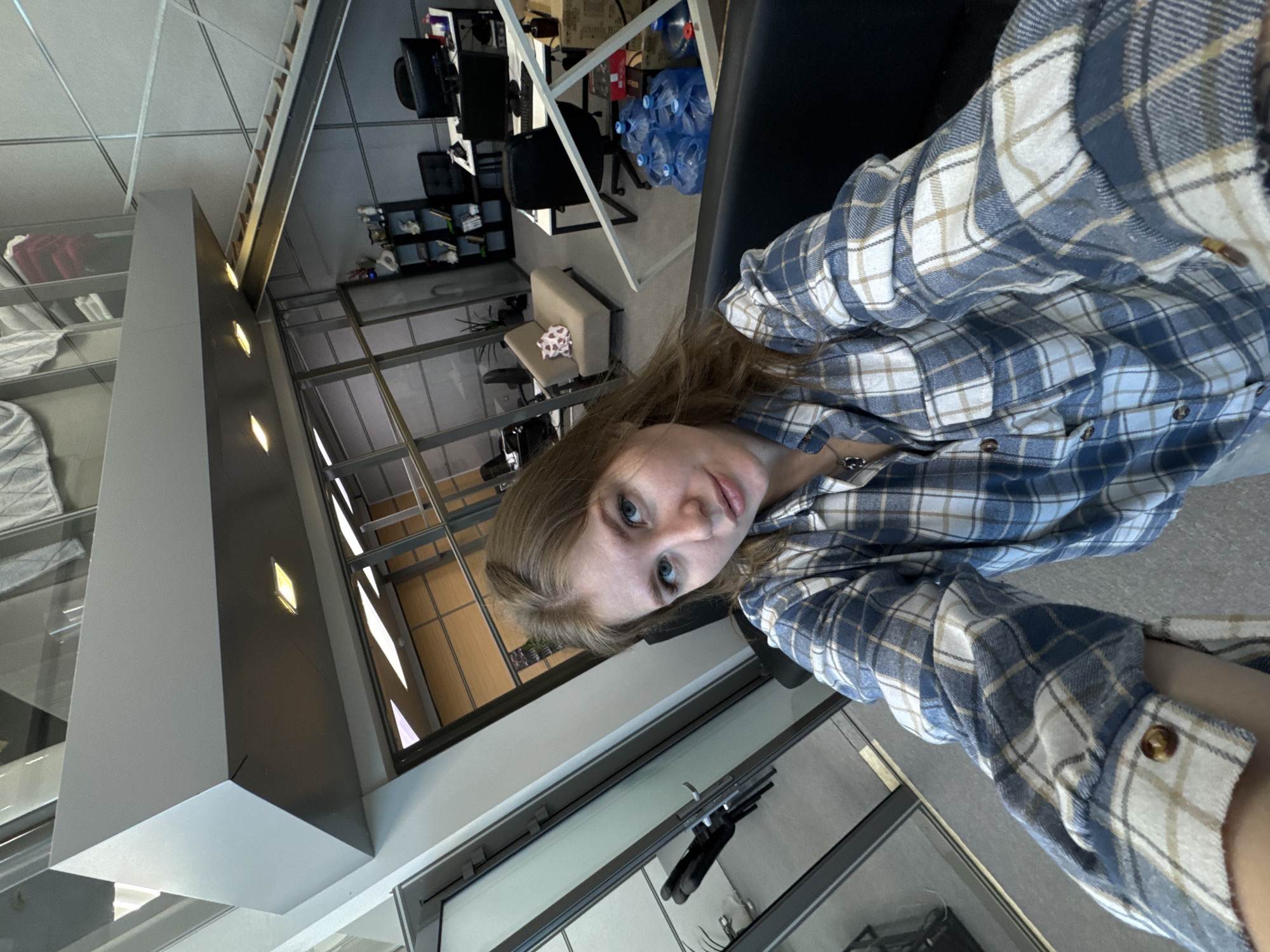 A person wearing a blue and white plaid shirt is seated in an office setting. Visible in the background are office chairs, desks, water bottles, and structural columns with integrated lighting. Through the window is a view, possibly of Perm, Russia, given the prompt.